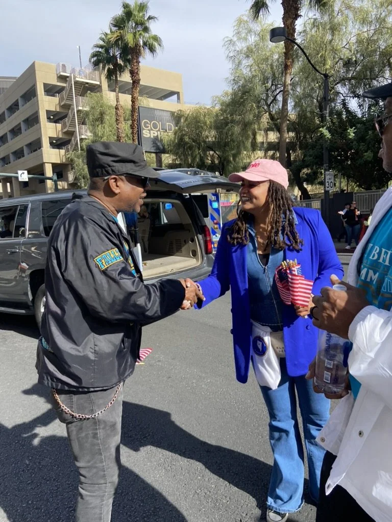 Two women shaking hands outdoors, one wearing a blue blazer and pink cap, the other in a gray jacket and black cap, while a third person stands nearby holding a water bottle. In the background, there are palm trees, a multi-story parking structure, and an emergency vehicle.
