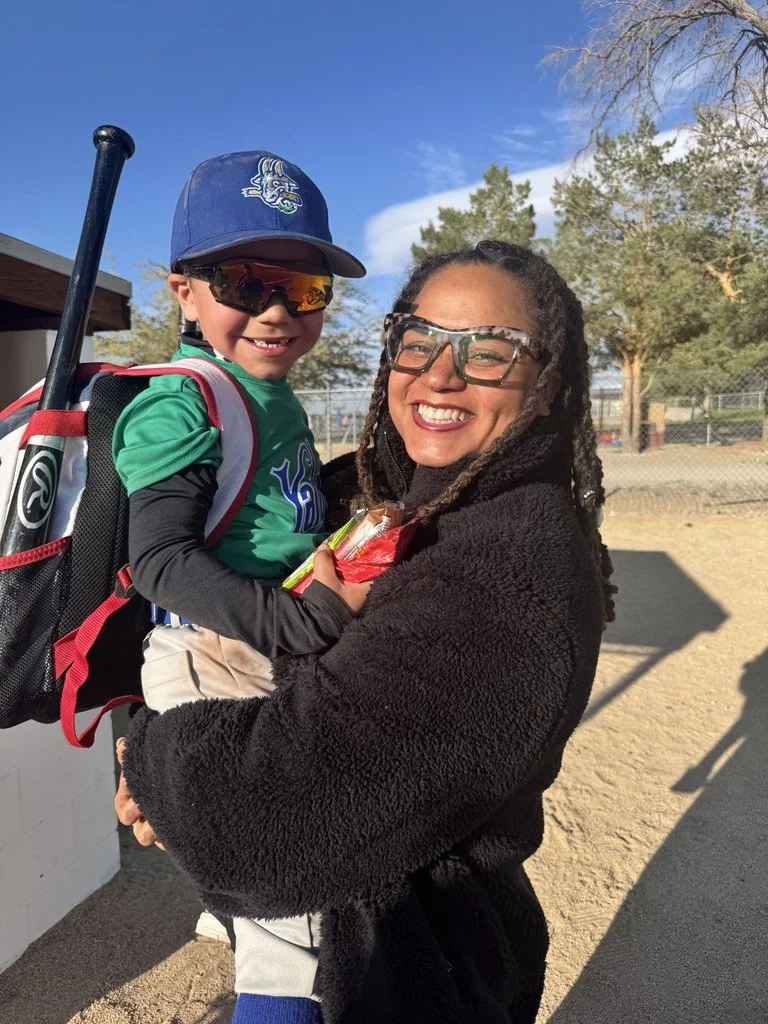 A woman holding a young boy at a baseball field on a sunny day. The boy is wearing a baseball uniform with a cap and sunglasses, holding a baseball bat. The woman is smiling, wearing glasses and a black jacket, with trees and a fence in the background.
