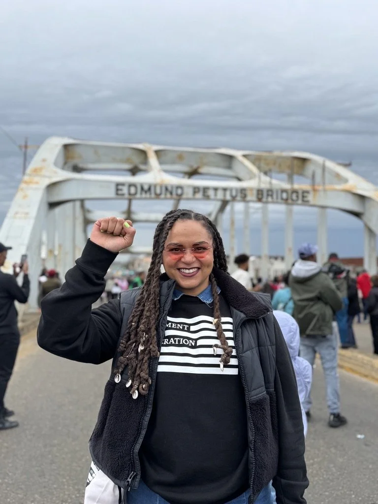 Haley Johnson, who is a smiling woman with braids and sunglasses, is raising her fist in front of the Edmond Pettus Bridge with voting rights activists and cloudy sky in the background.