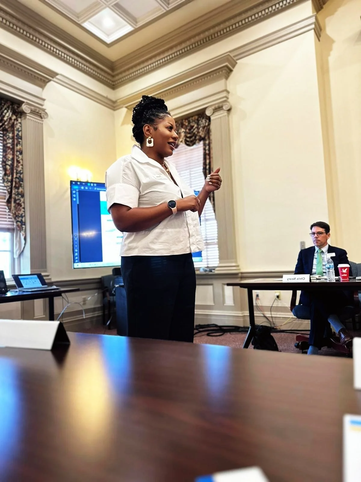 A woman standing and speaking at a conference or meeting, with a man seated at a table nearby, in a room with ornate walls and a large screen behind her.