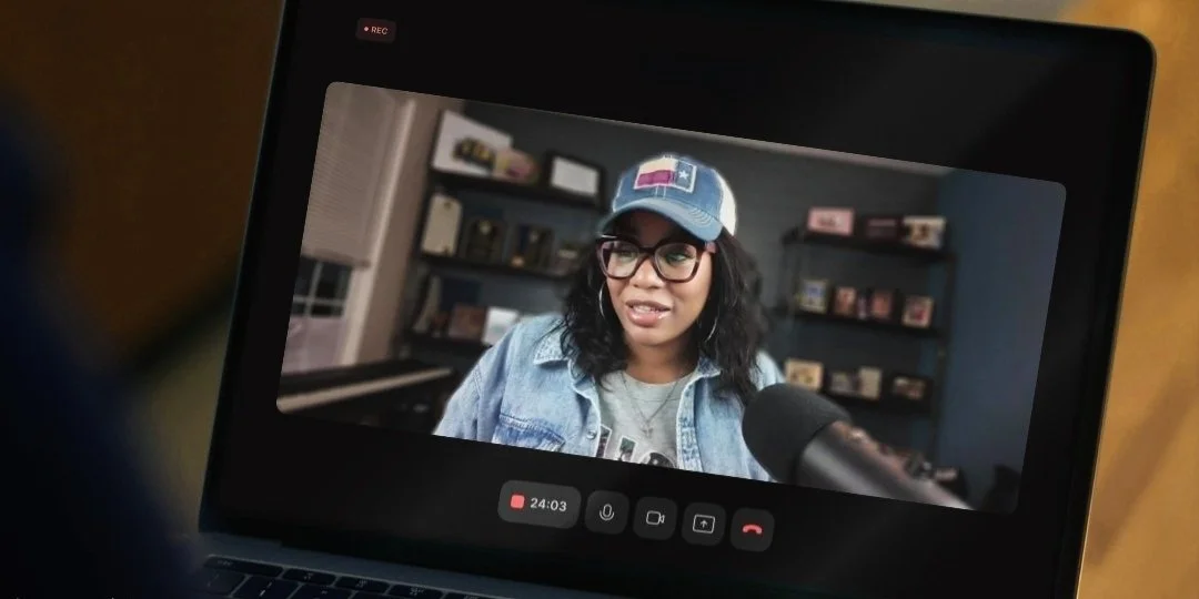 A woman wearing glasses, a denim jacket, and a cap is speaking into a microphone during a video call on a laptop screen.
