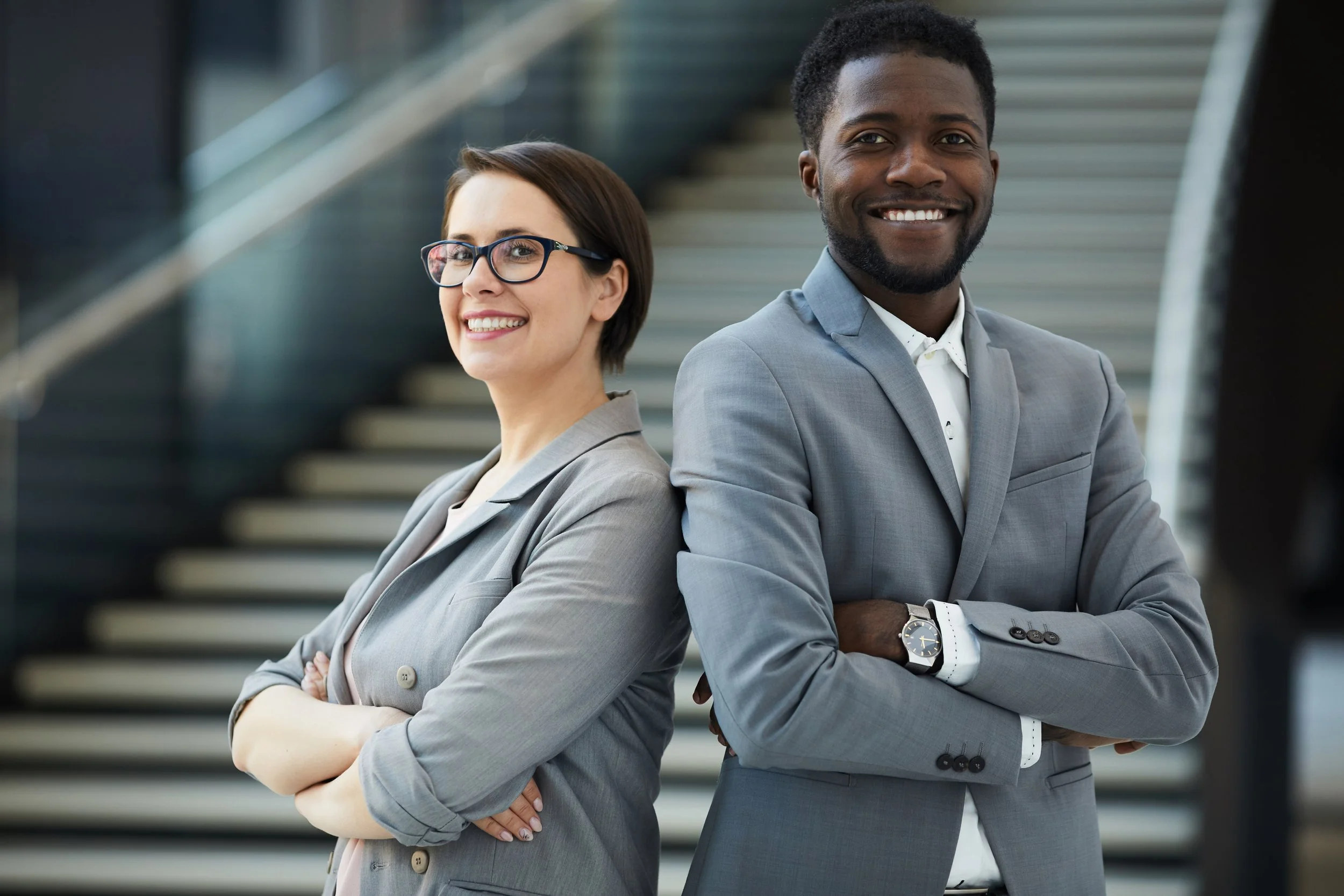 A woman and a man in professional attire standing back to back with arms crossed, smiling and looking at the camera on a staircase background.