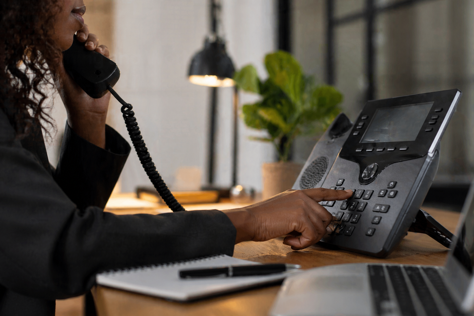 A woman with curly hair is speaking on a black landline phone while typing on a desk phone with a digital display and multiple buttons. There is a notebook with a pen on the wooden desk, with a laptop nearby and a potted plant in the background.