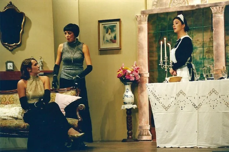 Tre donne in un soggiorno, una seduta su un divano e due in piedi, con una scena che sembra un mix tra un'opera teatrale e una scena cinematografica.