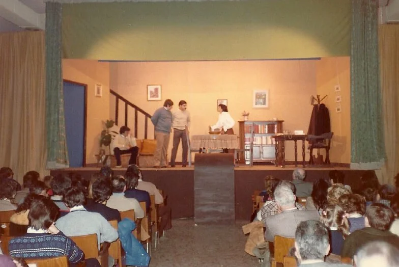 Scena teatrale con quattro attori sul palco, audience seduta che guarda, ambientazione domestica con mobili e decorazioni.