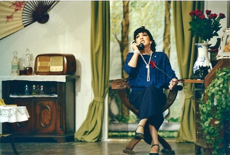 Una donna vestita elegantemente seduta su una sedia di legno, mentre parla al telefono in una stanza decorata con tende verdi, piante e oggetti d'antiquariato.