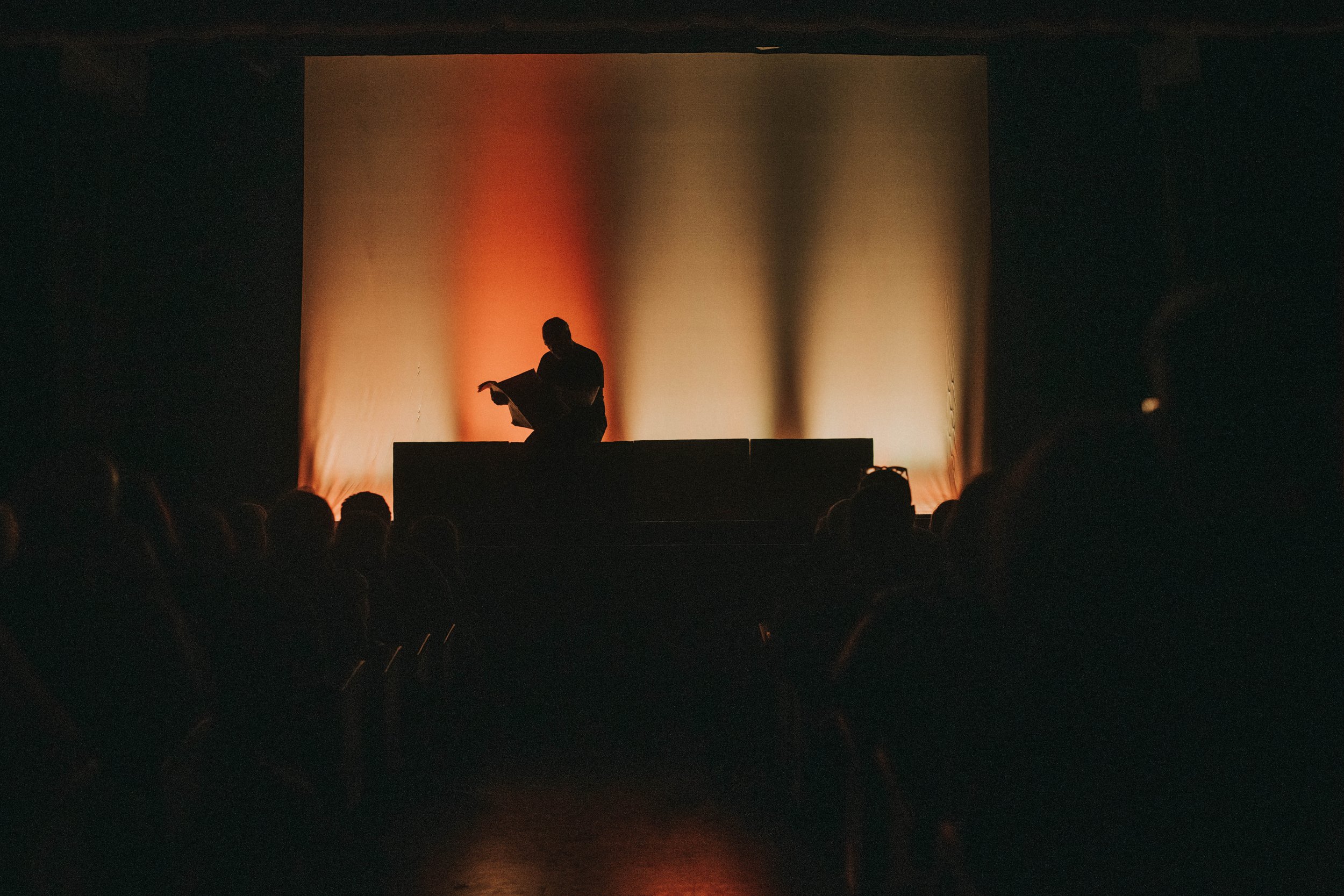 Una persona sul palco legge un libro davanti a un pubblico seduto, con una backdrop illuminata di colori caldi.