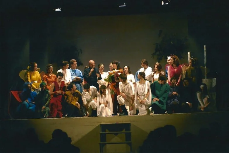 Gruppo di bambini e adulti su un palco durante uno spettacolo teatrale o una cerimonia, con alcuni bambini vestiti con costumi colorati e uno di loro che tiene un mazzo di fiori.