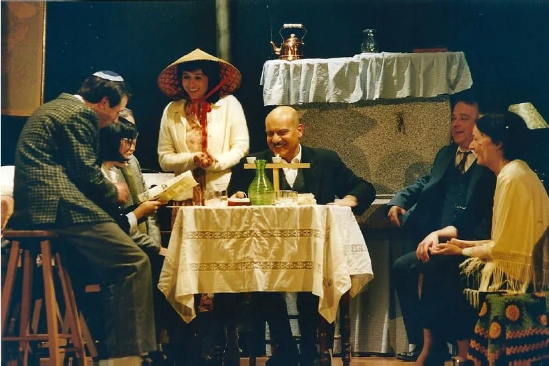 Un gruppo di persone durante uno spettacolo teatrale, sedute attorno a un tavolo con una tovaglia, alcune leggendo un programma, con un'ambientazione che richiama una scena di cucina vietnamita.