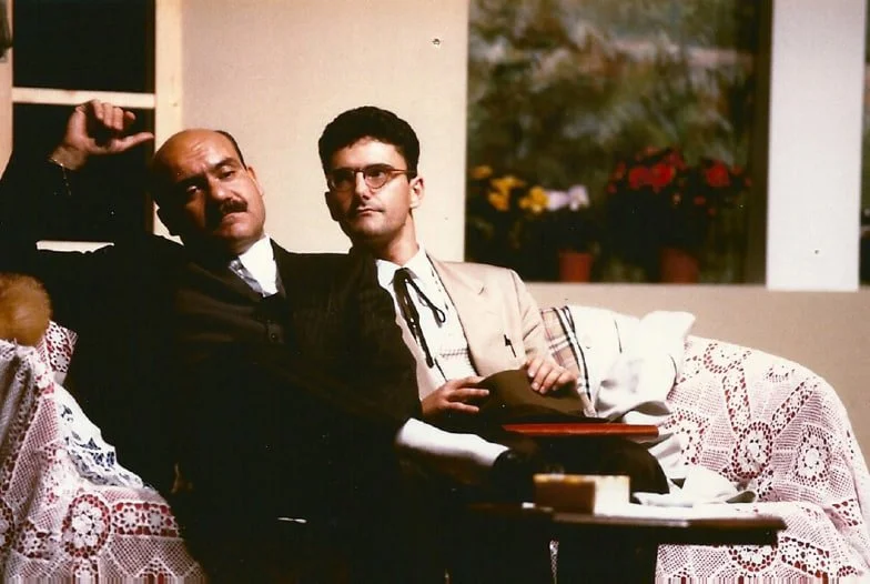 Due uomini seduti su un divano, uno più anziano con barba e cappello, l'altro più giovane con occhiali, in un soggiorno con una finestra e piante sullo sfondo.