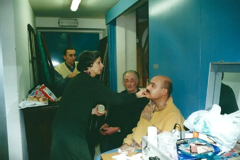 Una scena di una sessione di trucco o trucco su un uomo seduto a una scrivania, con altre tre persone intorno, in un ambiente interno con pareti blu e oggetti vari sulla scrivania.