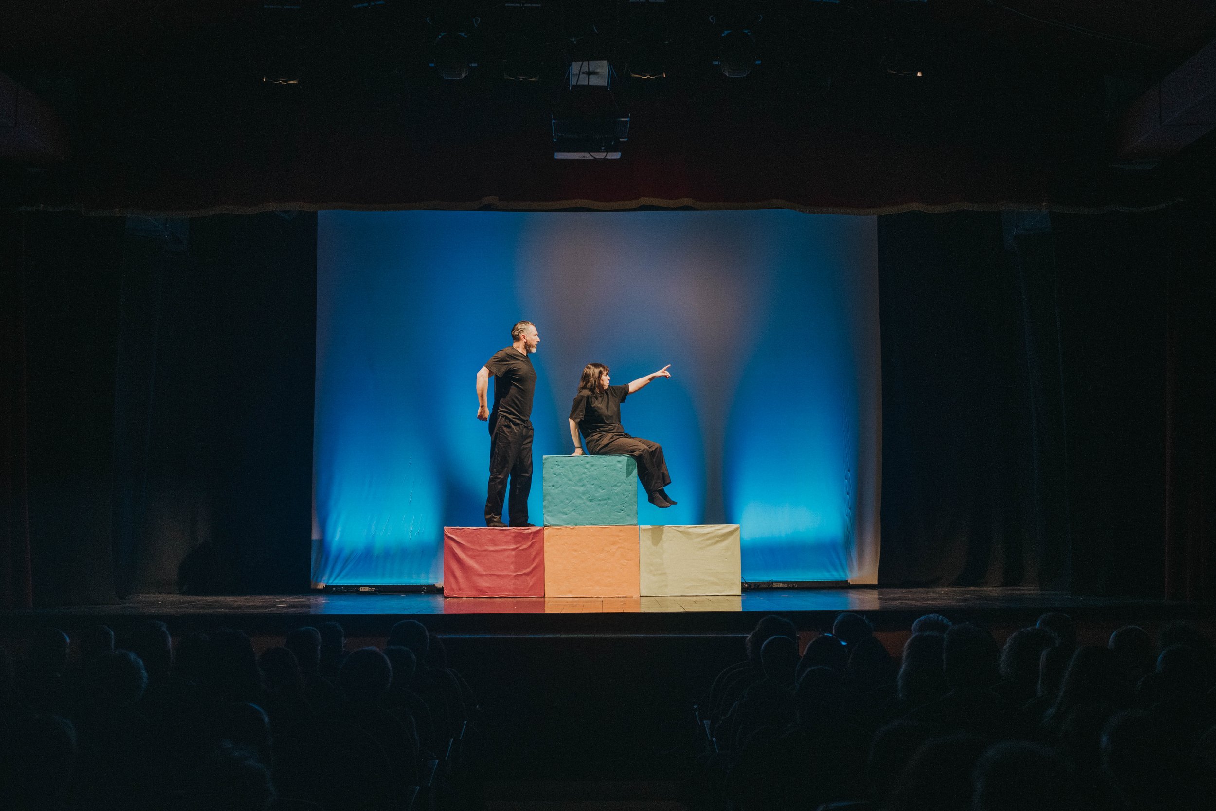 Due persone stanno facendo uno spettacolo di teatro su un palcoscenico con blocchi colorati, uno dei quali è seduta e l'altra in piedi accanto