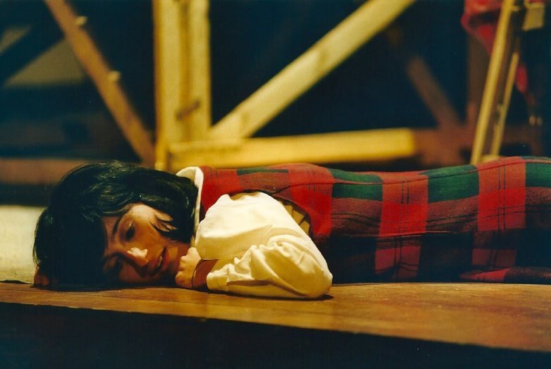 Una donna con capelli neri corti, che si trova sdraiata sul pavimento di legno, con un sorriso, vestita con una camicia bianca e un gilet a scacchi rossi e verdi.