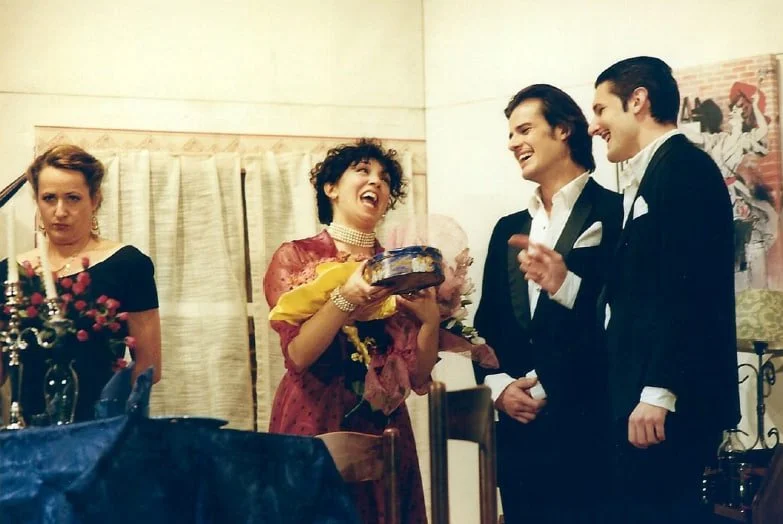 Due uomini in tuxedo sorridono e parlano con una donna con un vestito rosso e collana, mentre un'altra donna in abito nero si guarda sconsolata.