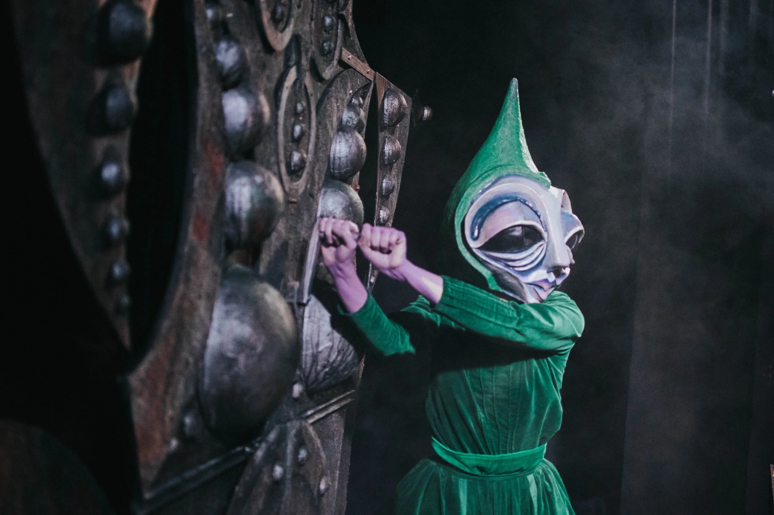 Persona con maschera verde e vestito verde che rompe una grande maschera di legno.