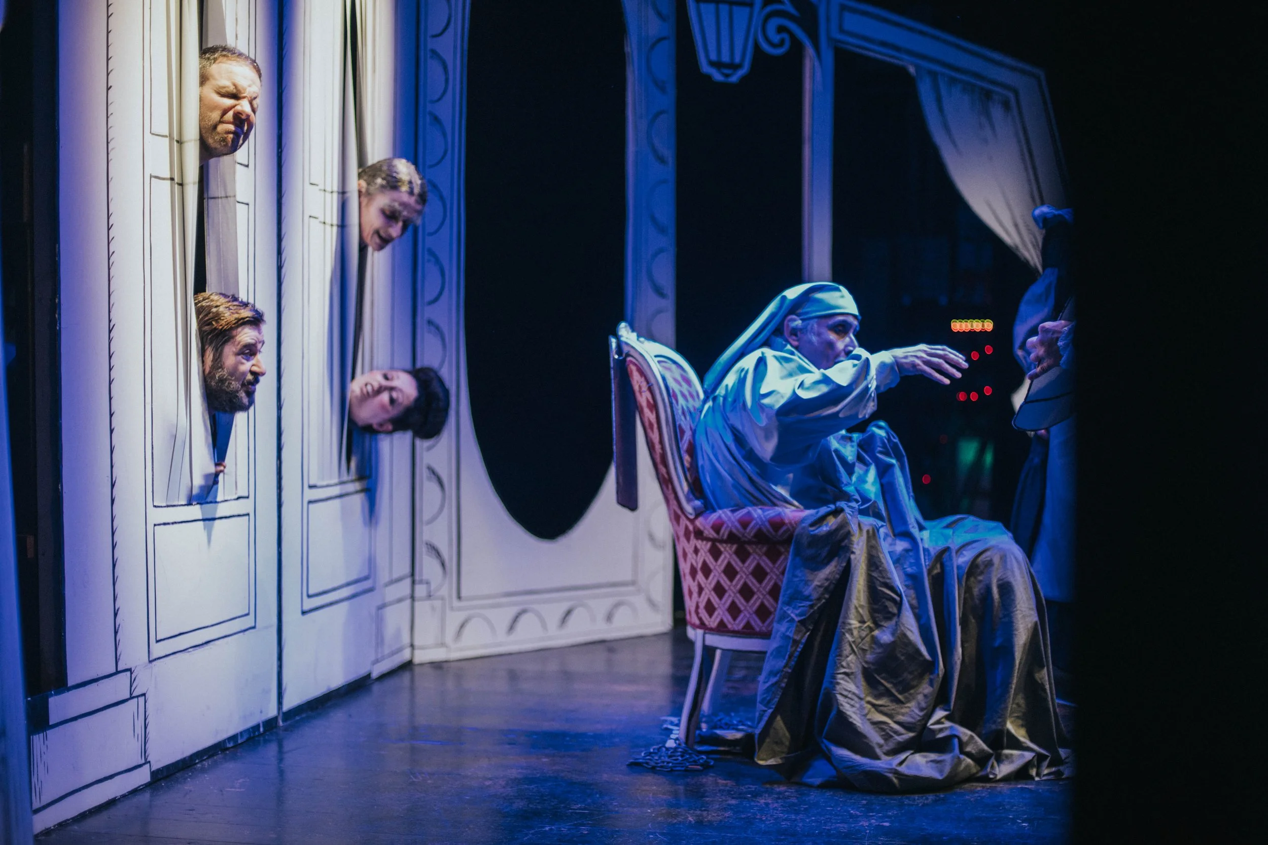 Scena teatrale con personaggi in costume, uno seduto su una sedia e altri che sporgono da un set disegnato con pannelli e un quadro, in un teatro con luci blu e neri.