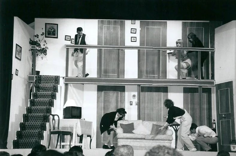 Scena teatrale con cinque persone, due sulla soglia del secondo piano, due attorno a un divano in primo piano, una vicino alla scala, in una stanza con mobili e decorazioni.