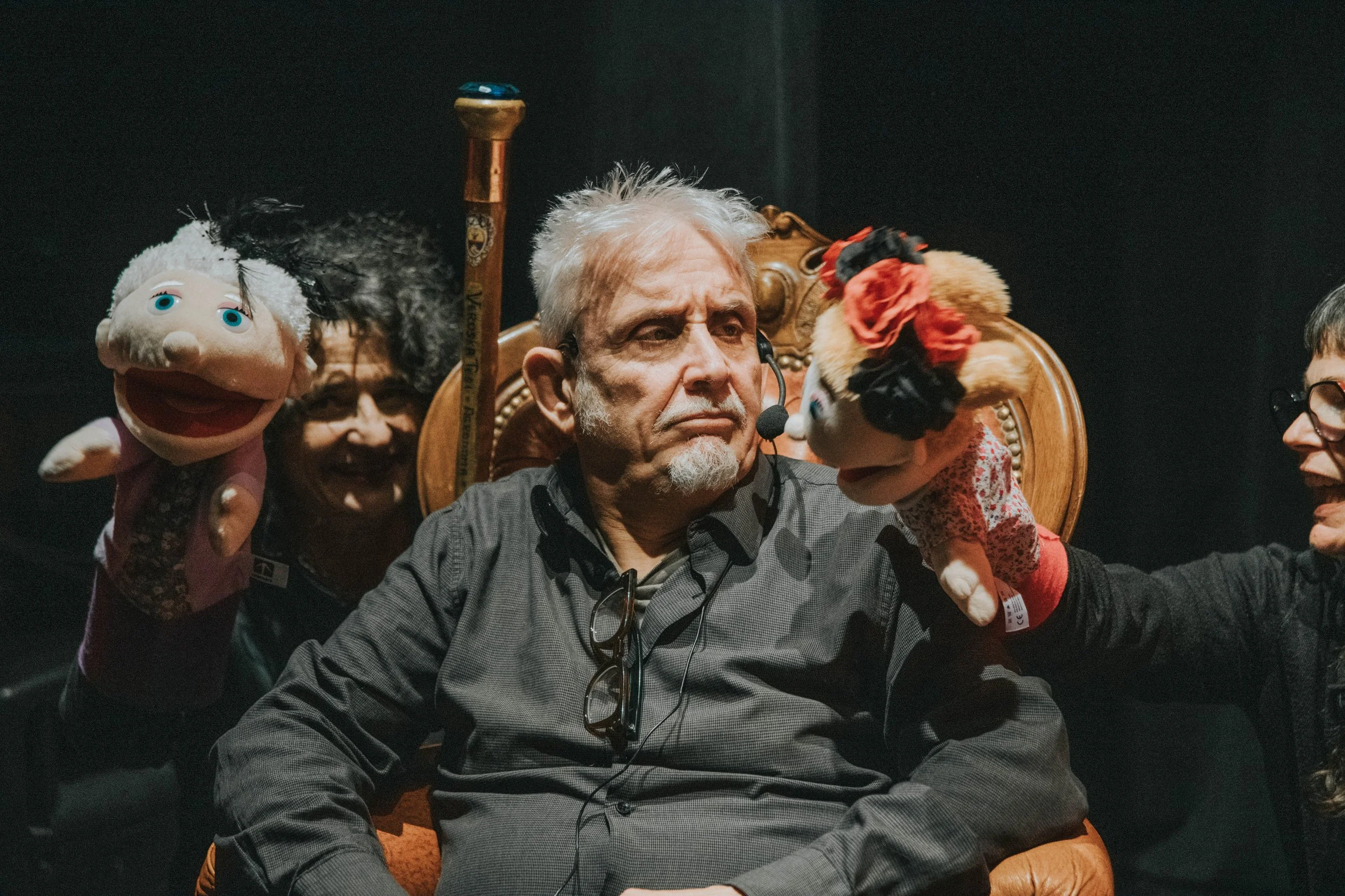 Un uomo anziano con cuffie e microfono seduto su una sedia di legno antidella, circondato da burattini con figurini umani, in uno spettacolo di marionette.