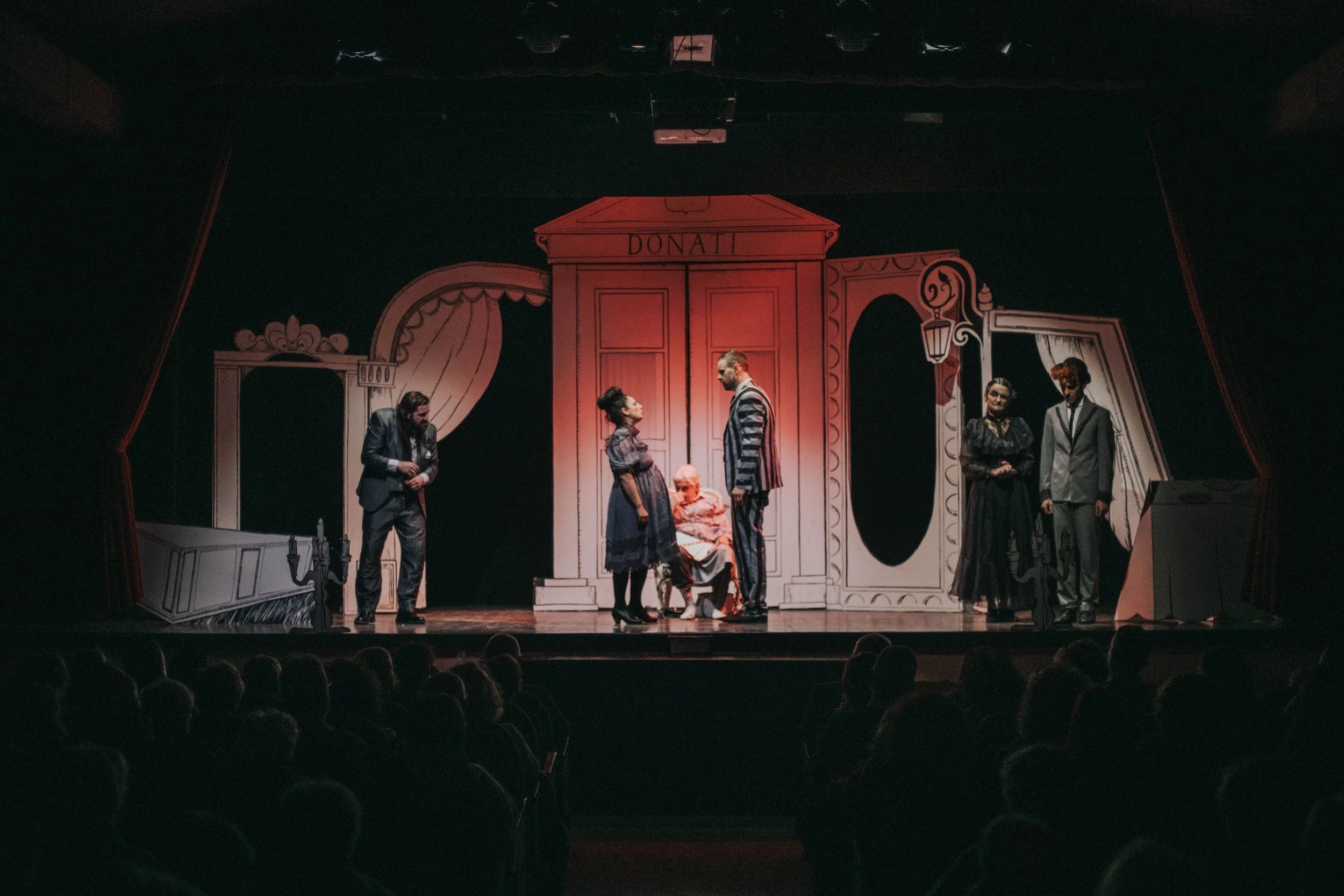 Scene di una recita teatrale con attori in costume su un palcoscenico decorato con uno sfondo disegnato di un ambiente interno, con un gruppo di persone che recitano in una scena che coinvolge un'anziana donna seduta e altri personaggi che interagisc