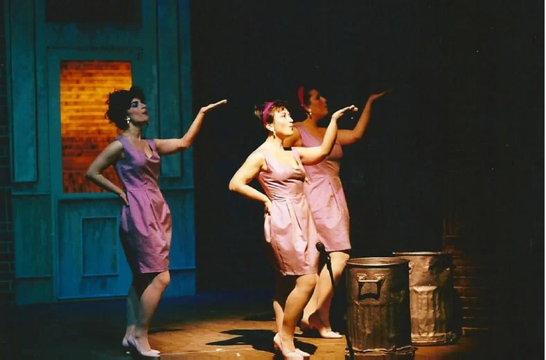 Tre donne in abiti rosa si esibiscono in una scena teatrale, con due bidoni sul palco.