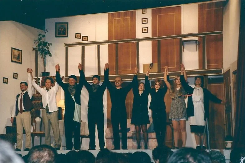 Gruppo di persone che si inchinano sul palco di uno spettacolo o una recita, con le mani alzate insieme in segno di vittoria o ringraziamento, in un ambiente teatrale.
