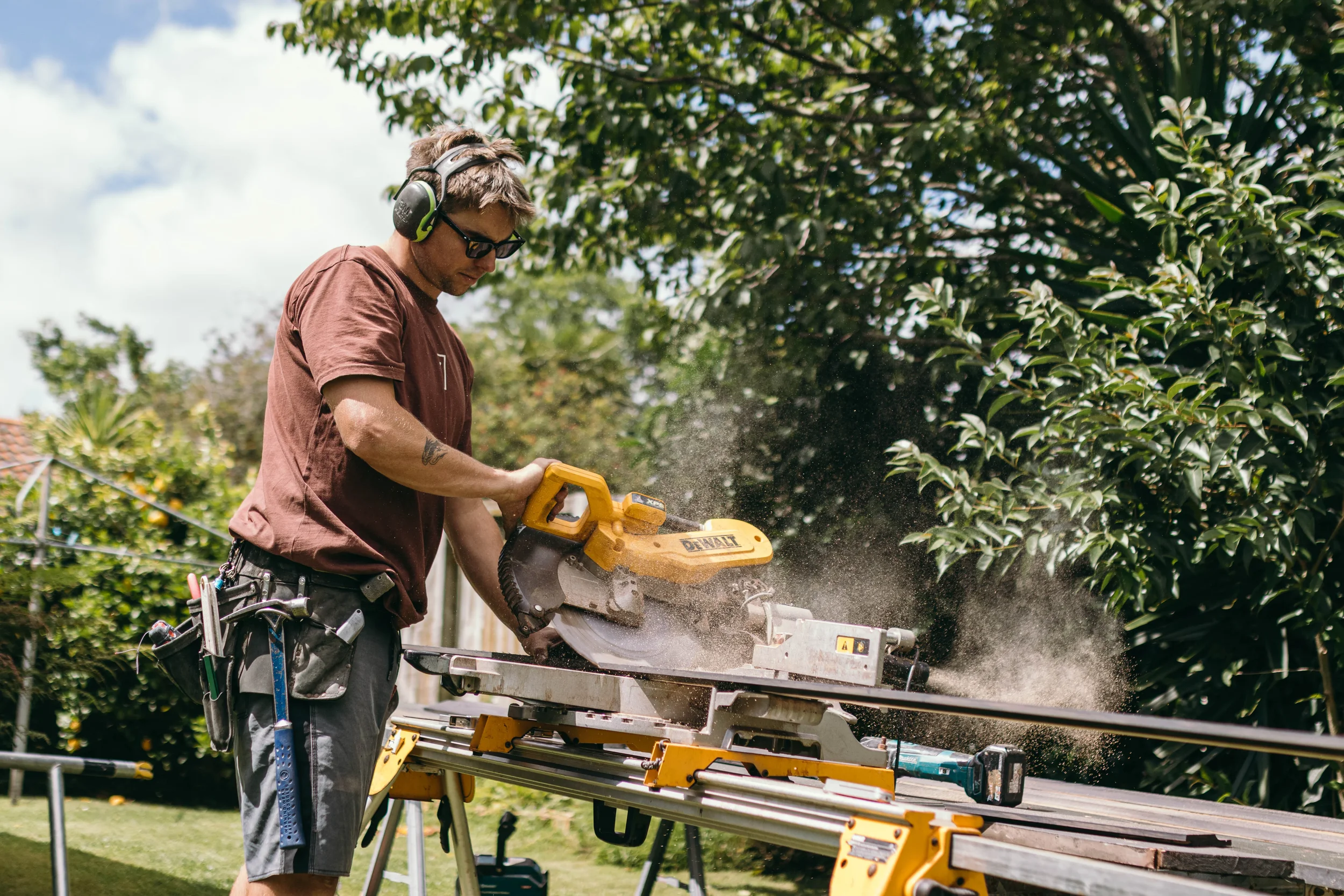 Builder from Zephyr Construction cutting wood with a yellow sliding miter saw outdoors, wearing safety glasses and hearing protection.