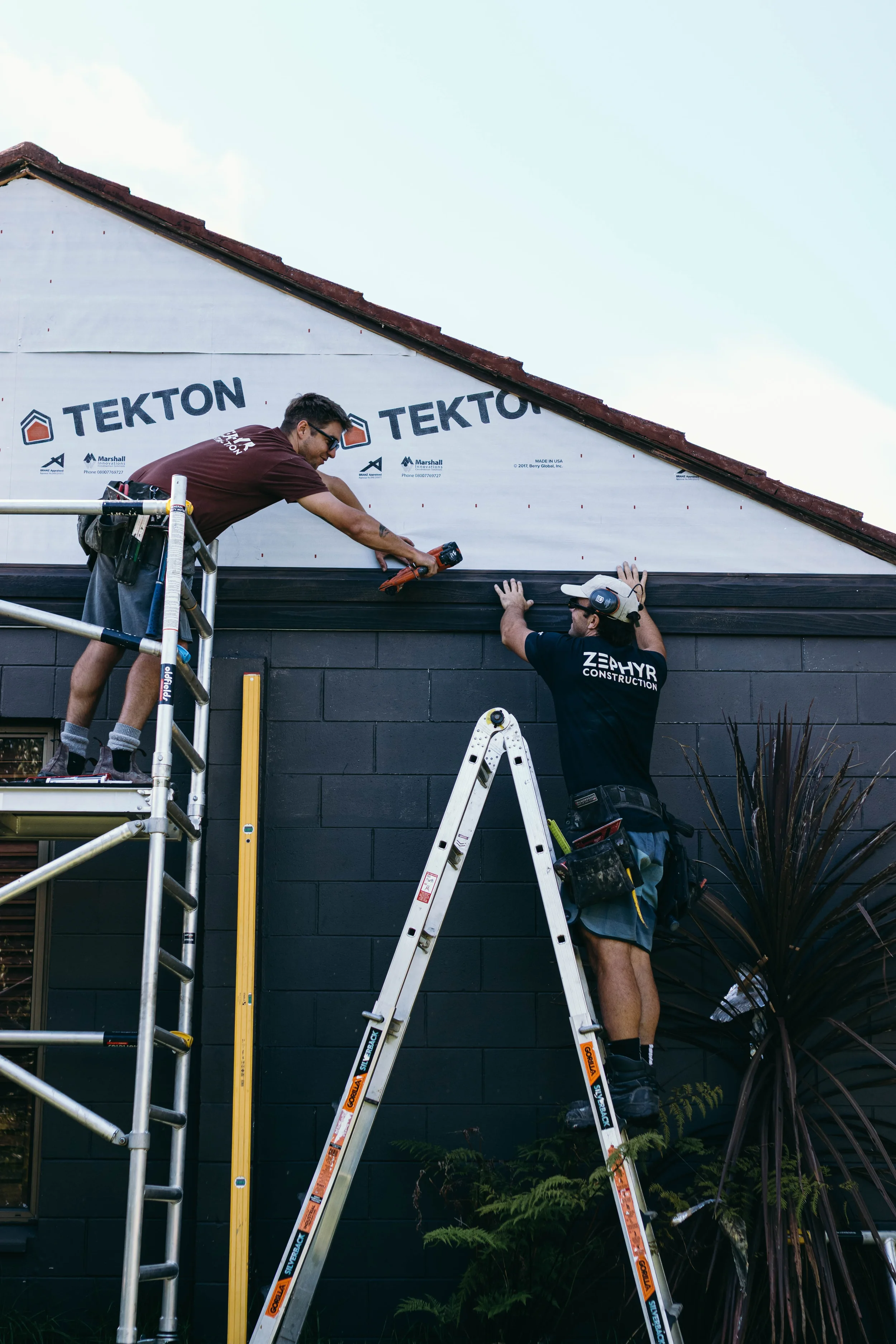 Zephyr Construction team installing cedar cladding on a house exterior. One worker is standing on a scaffold, passing tools to another worker on a ladder, who is securing the siding near the roof.