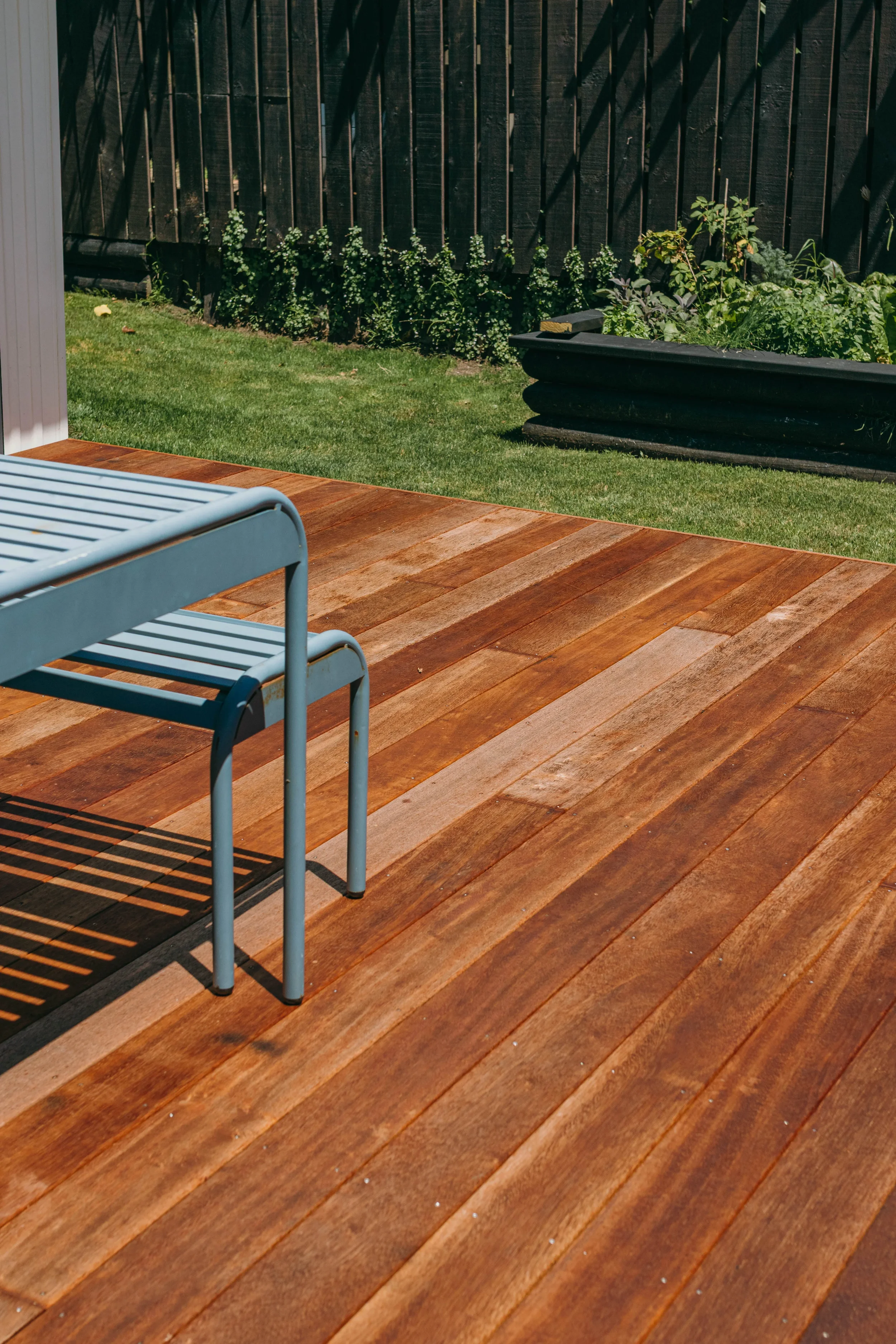 A backyard in the Bay of Plenty with a wooden deck built by Zephyr Builders, metal chairs, a grassy area, and a raised garden bed with plants against a dark wooden fence.