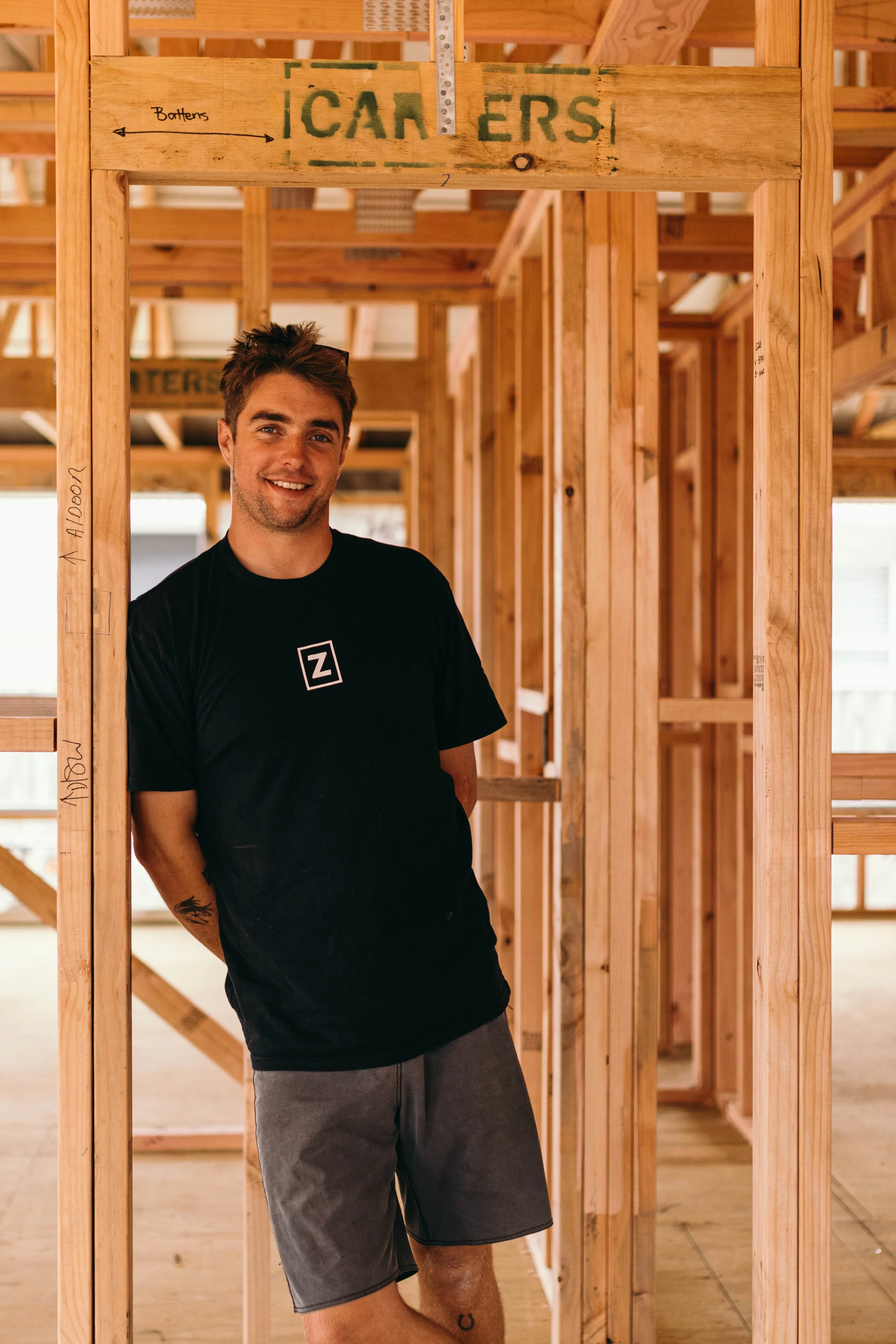 Jack Jellyman from Zephyr Construction stands among the wooden framing of a house under construction, wearing a black t-shirt and gray shorts.