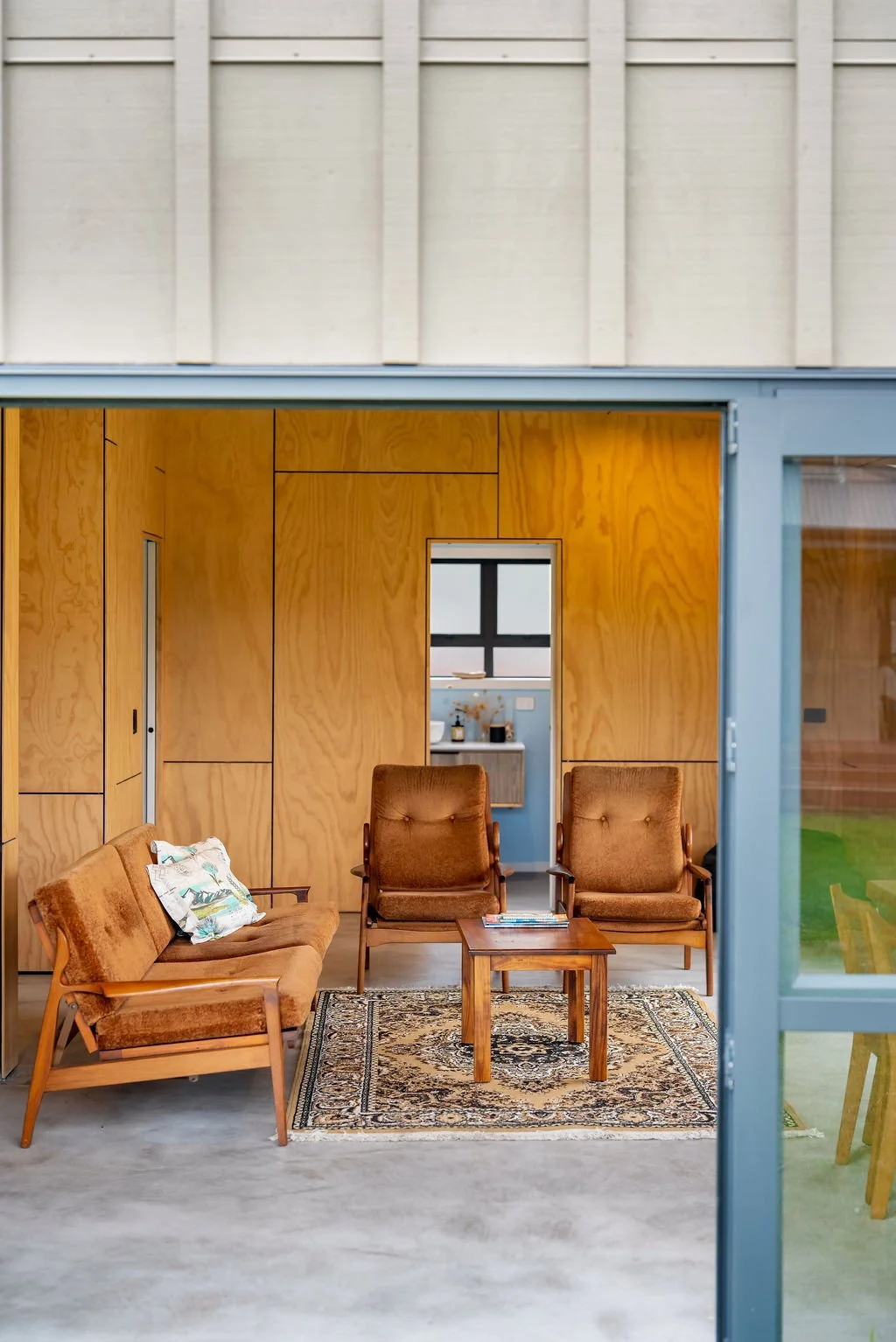 View of a cozy seating area inside a modern room with wooden walls, containing vintage-style brown armchairs, a small wooden table, and a patterned rug, seen through an open glass door.
