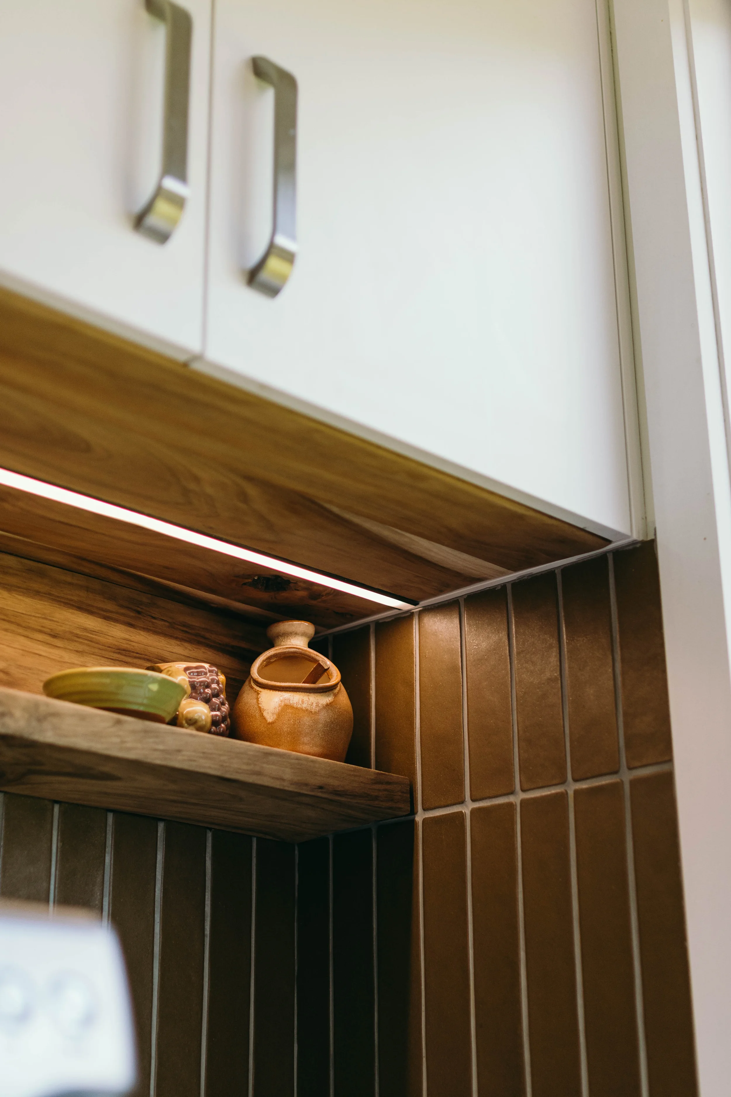 Kitchen corner with white cabinets, wooden shelf holding ceramic dishes and a small ceramic jug, wabi-sabi brown tiled backsplash renovated by Zephyr Construction.