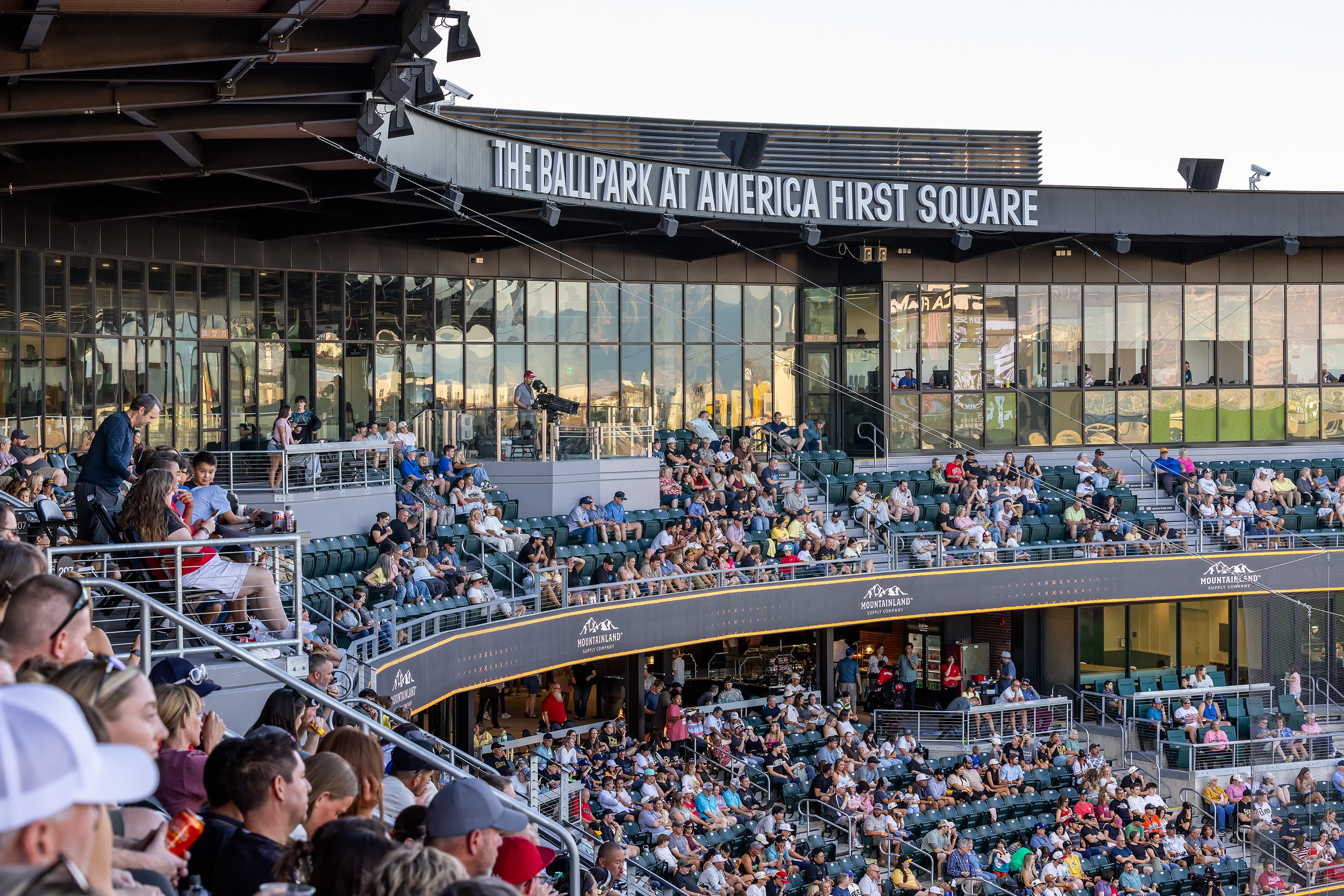 Crowd seated in outdoor baseball stadium at America First Square, with glass-fronted building in background.