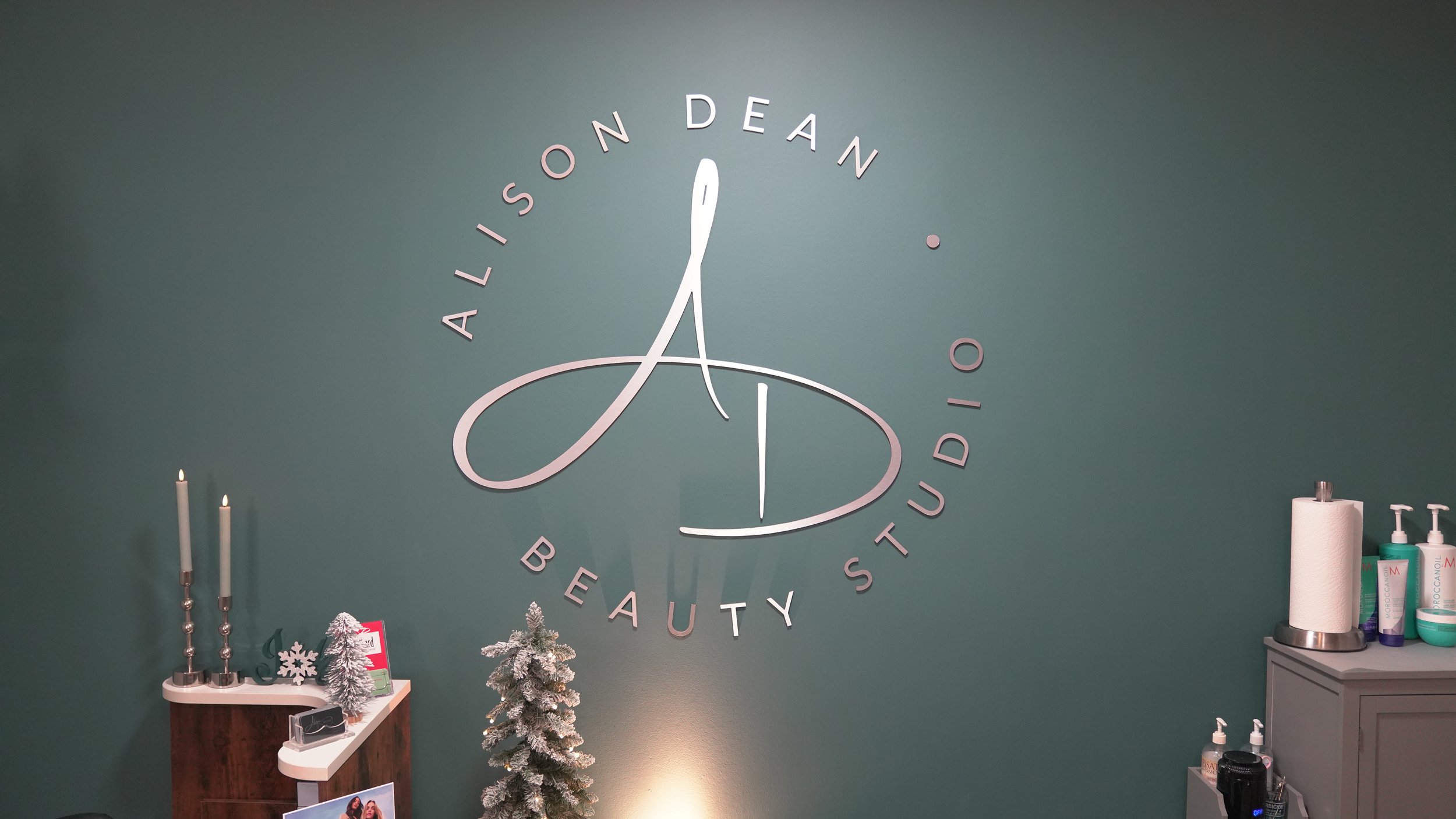 Decorated beauty studio wall with large logo, two lit candles, small artificial Christmas tree, and various self-care products on counter.