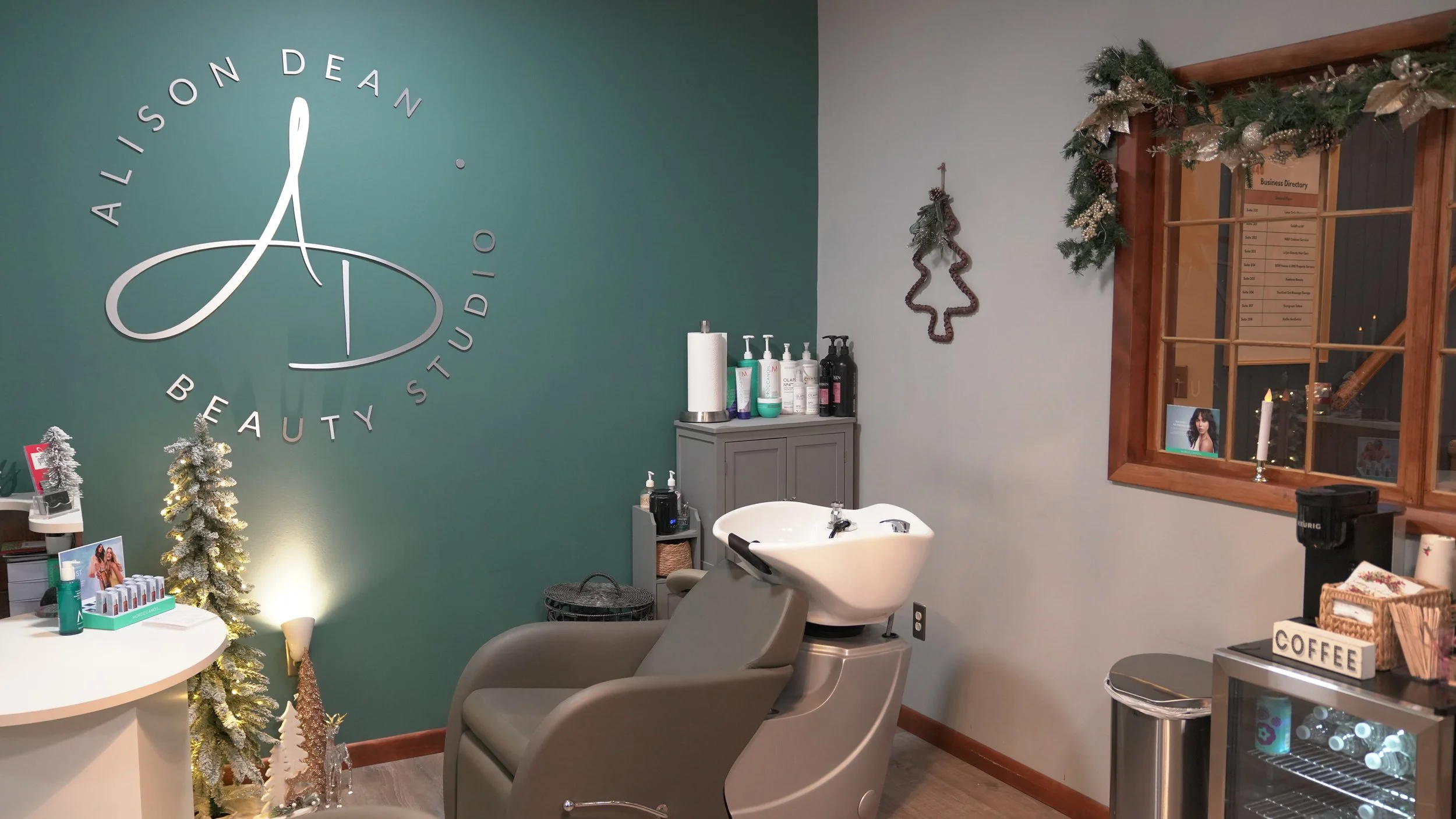 Interior of a beauty studio decorated for Christmas, with a teal wall displaying the logo 'Alison Dean Beauty Studio', a small Christmas tree, and a salon chair with a wash basin. There is a window with holiday garland and a coffee station.