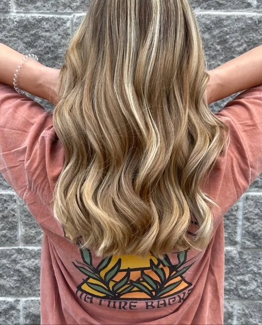 Back of a woman with long, wavy blonde hair wearing a pink t-shirt with a graphic of a sunset and leaves on the back, standing against a gray brick wall.