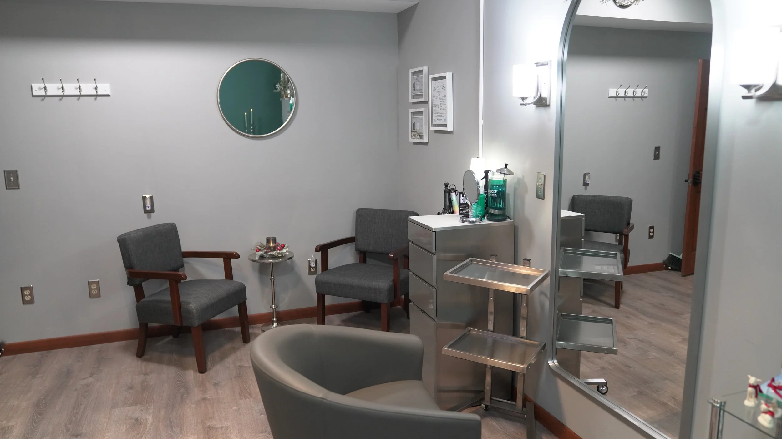 A room with gray walls and wooden trim, furnished with two gray armchairs, a small metallic side table, and a large mirror, and decorated with framed pictures and hooks on the wall. A plastic cart with drinks and a water dispenser sits next to a wall mirror. The room has a wooden floor and bright lighting.