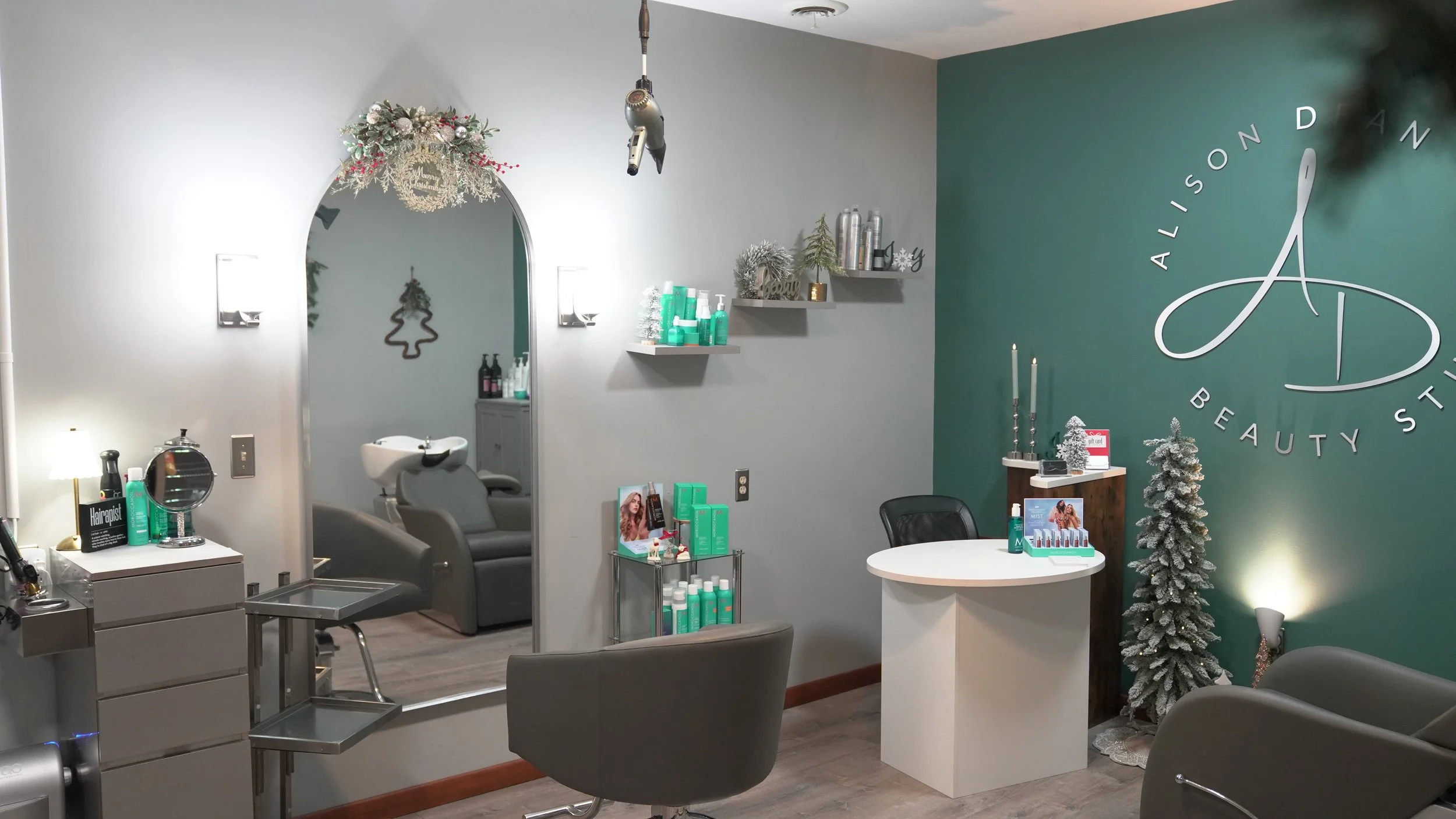 Interior of a beauty salon with a large green wall featuring the salon's logo, decorated for the holidays with a snow-dusted Christmas tree, candles, and holiday decor, along with salon chairs, shelves with hair products, and a reception area.
