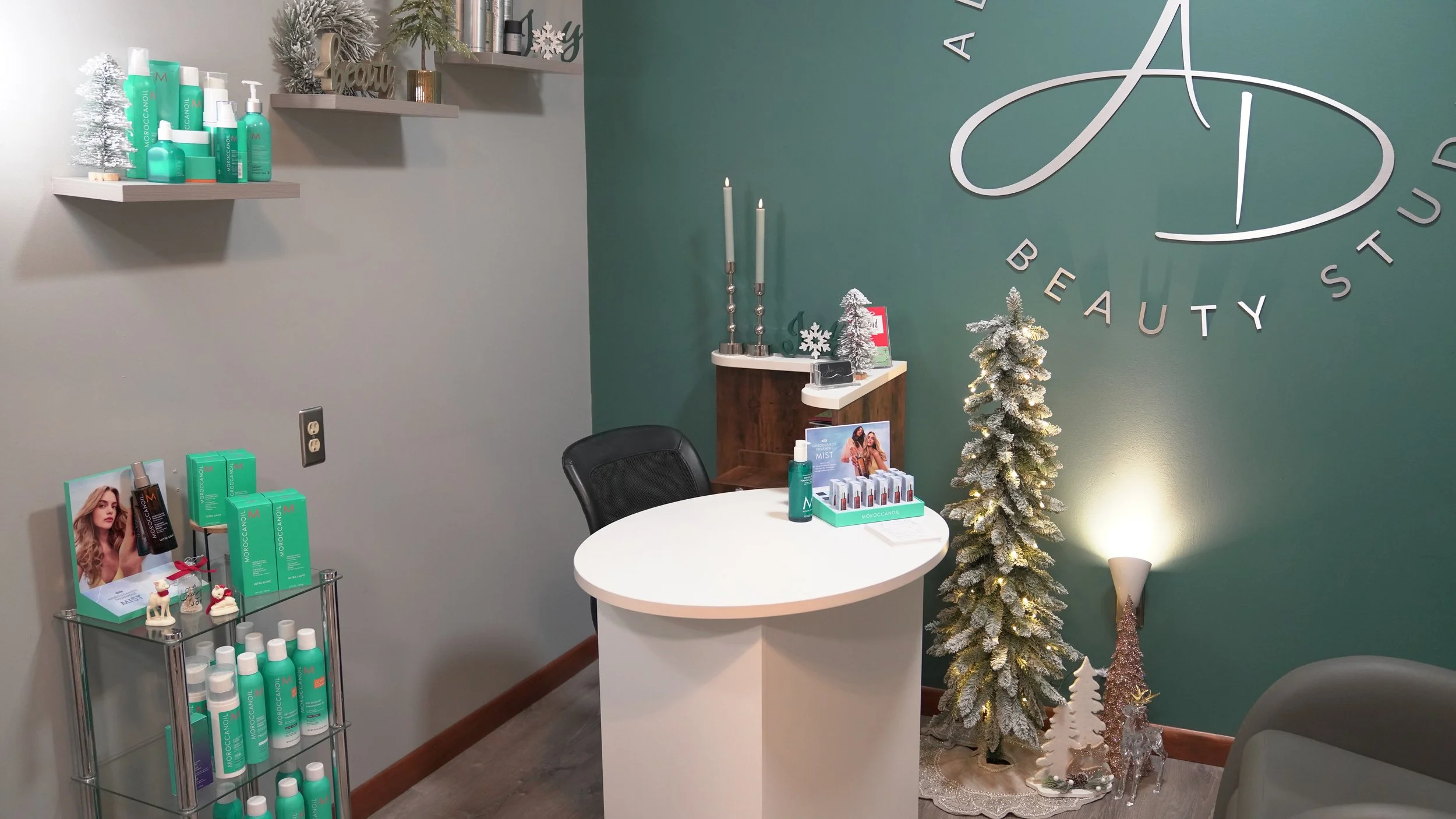 Interior of a beauty studio decorated for the holidays with Christmas trees, candles, and beauty products on shelves and display stands, featuring a white reception desk and a teal accent wall with a large logo.