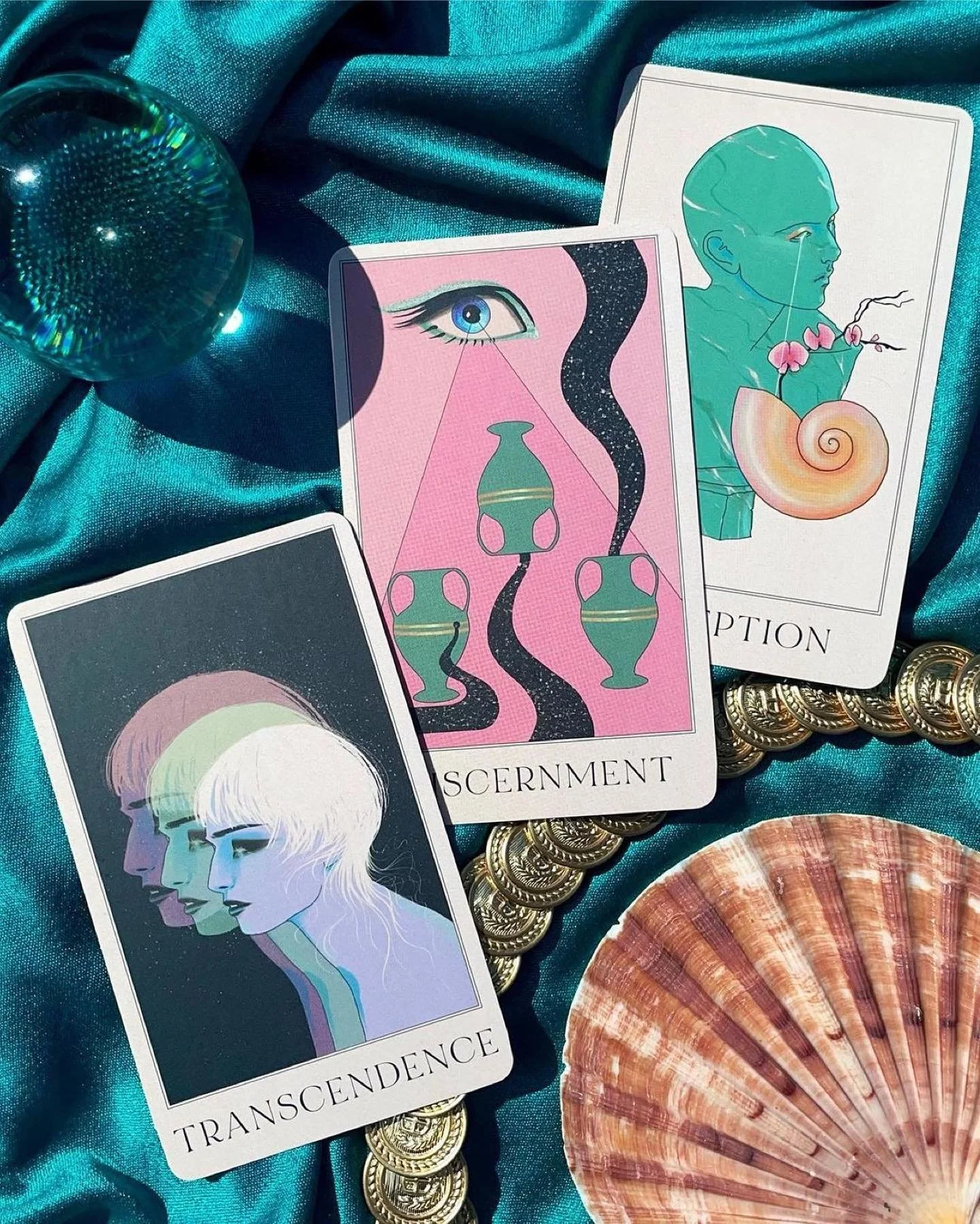 Three oracle cards from the Modern Nirvana deck displayed on a teal satin cloth, with a seashell, gold coins, and a small blue glass orb nearby.