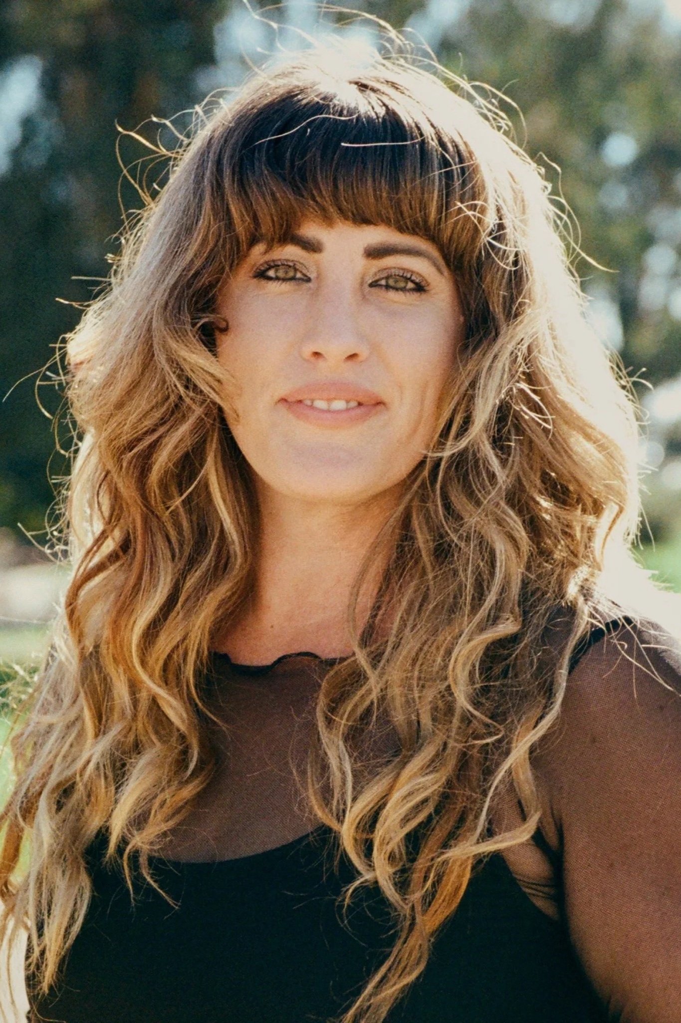A woman with long, wavy brown hair and bangs, smiling outdoors on a sunny day, wearing a black top.