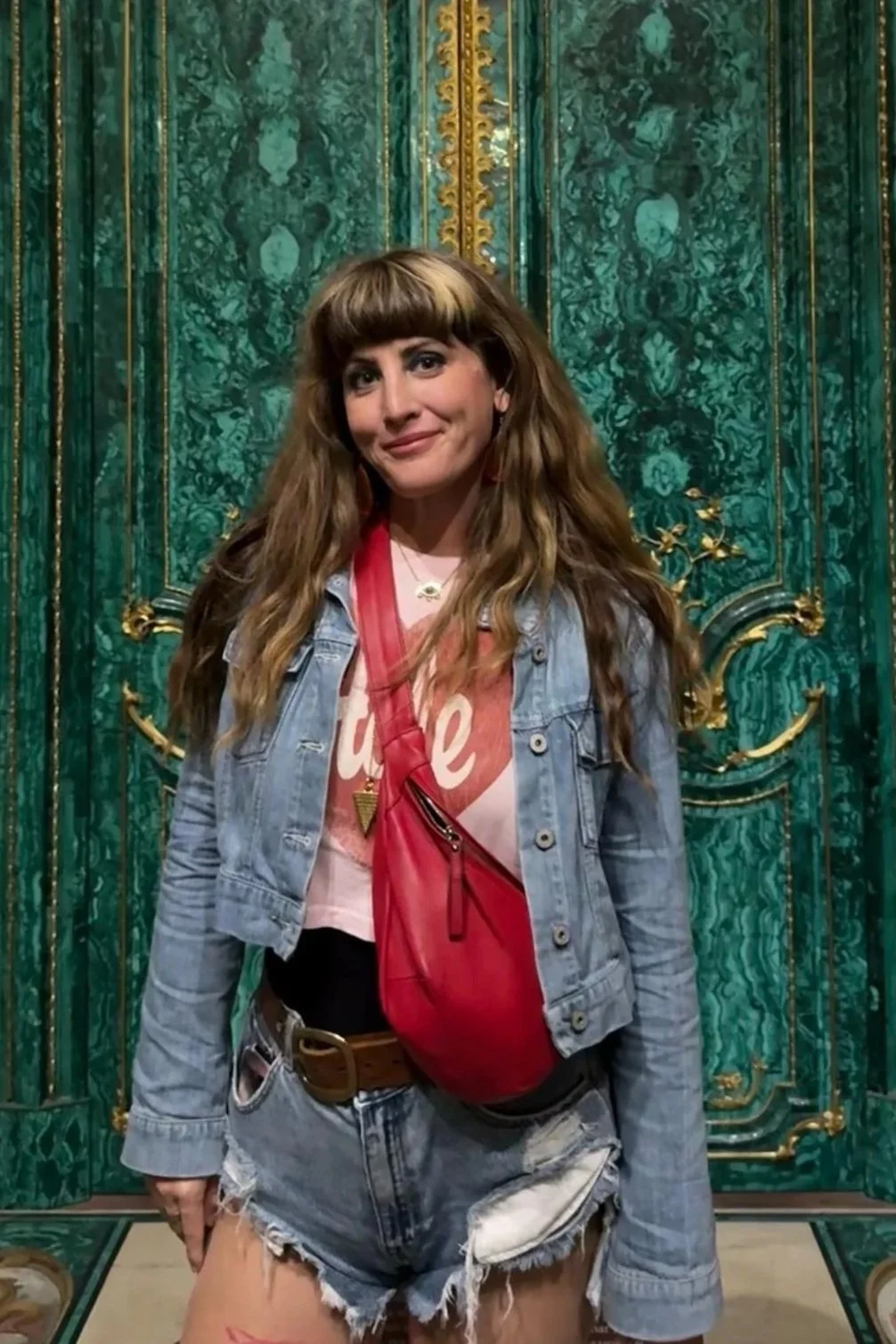A woman with long wavy hair and bangs standing in front of an ornate green and gold decorative wall. She is wearing a pink graphic T-shirt, a light blue denim jacket, and distressed denim shorts. She has a pink crossbody bag and a necklace with a small pendant.