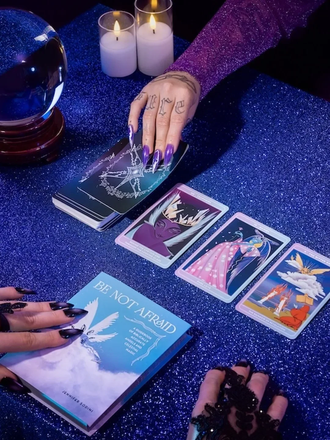 A person with purple glittery clothing and long purple nails holding tarot cards on a dark blue glittery cloth, surrounded by candles and a crystal ball, with a blue book titled 'Be Not Afraid' nearby.