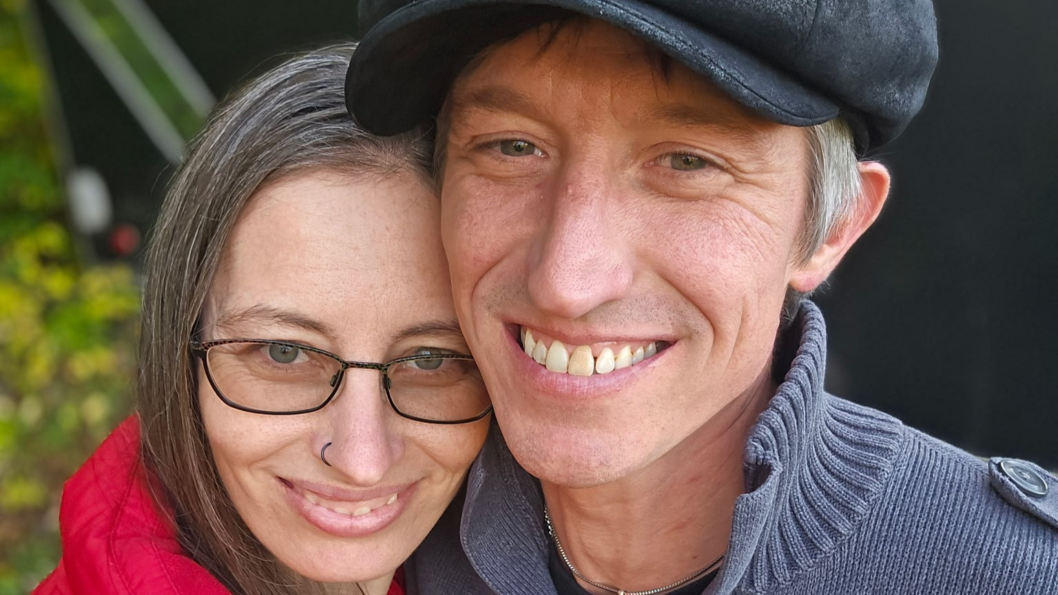 Close-up of a smiling couple embracing outdoors; woman has long brown hair, glasses, and a nose ring, wearing a red jacket; man has short hair, wears a black cap and a gray sweater.