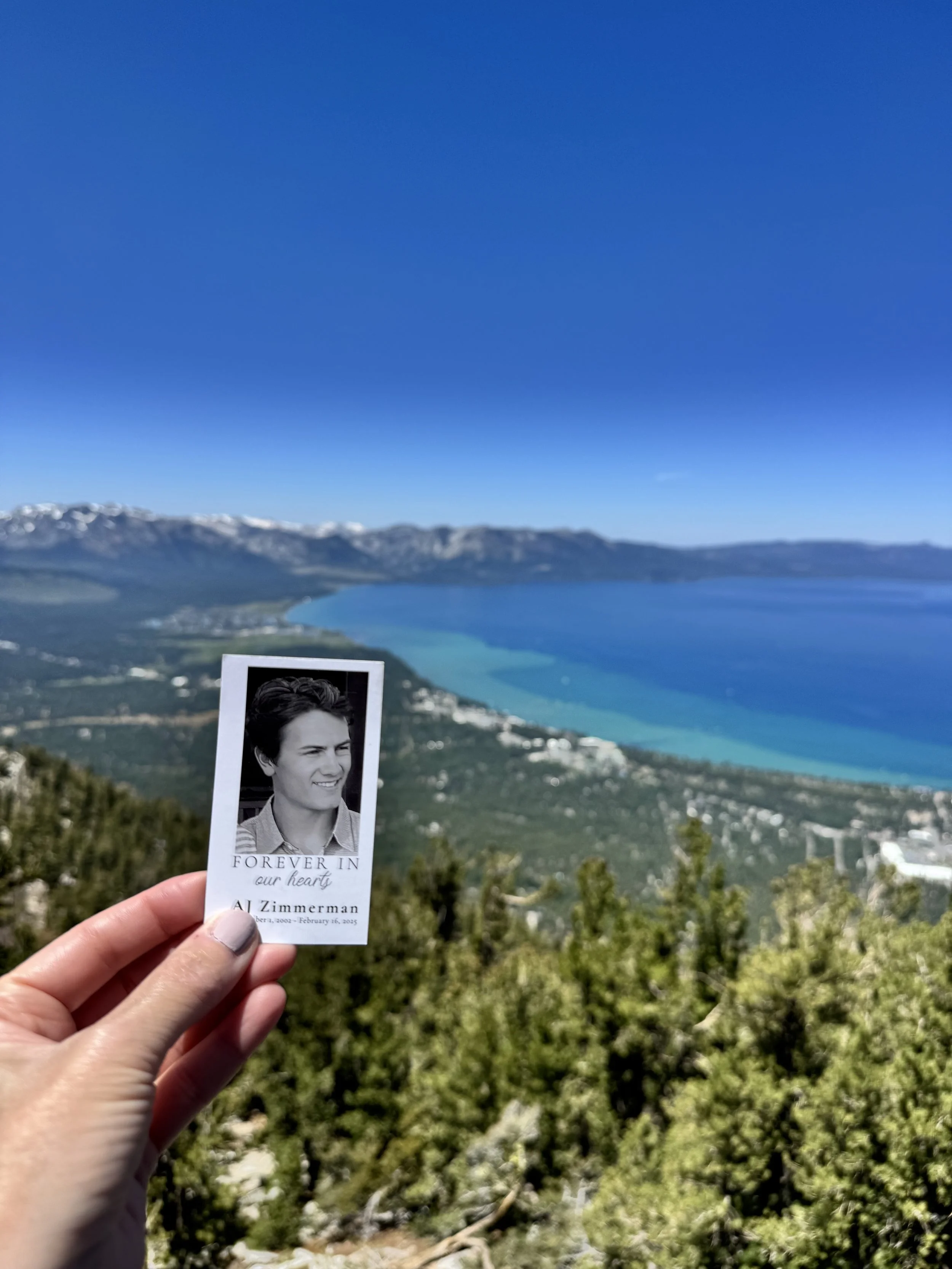 Person holding a memorial card with a photograph and text in front of a scenic view of a lake, mountains, and trees.