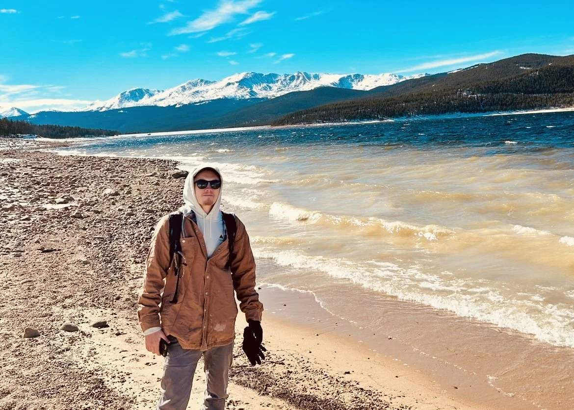 A man dressed in outdoor winter clothing, wearing sunglasses, gloves, and a backpack, standing on a sandy beach near a large body of water with snow-capped mountains in the background under a clear blue sky.