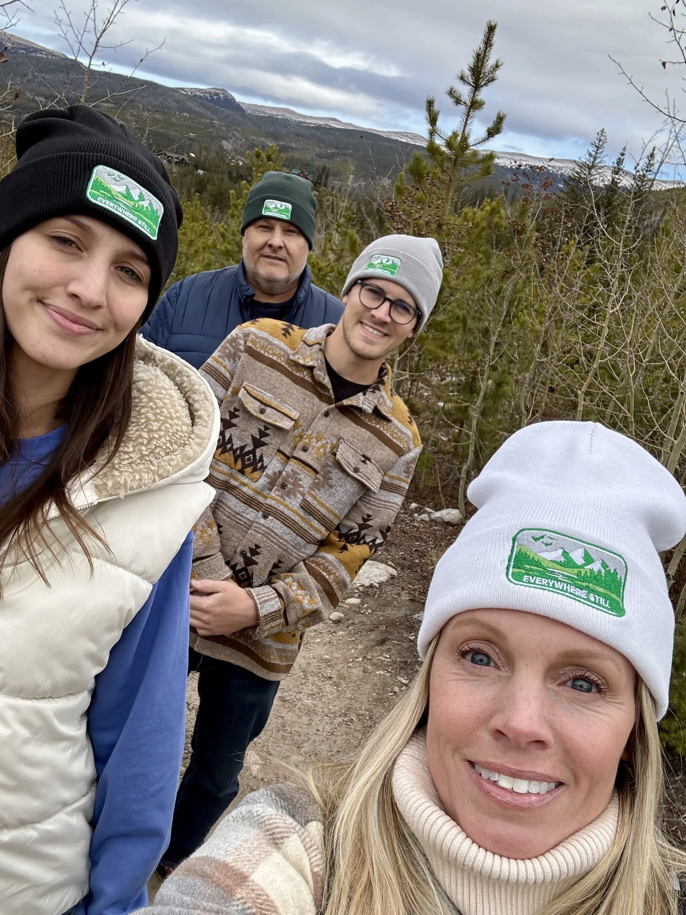 Four people outdoors on a trail in a mountainous, forested area, wearing warm clothing and beanies with a green mountain logo and the words "Everywhere Still." The background shows trees and snow-capped mountains under a cloudy sky.