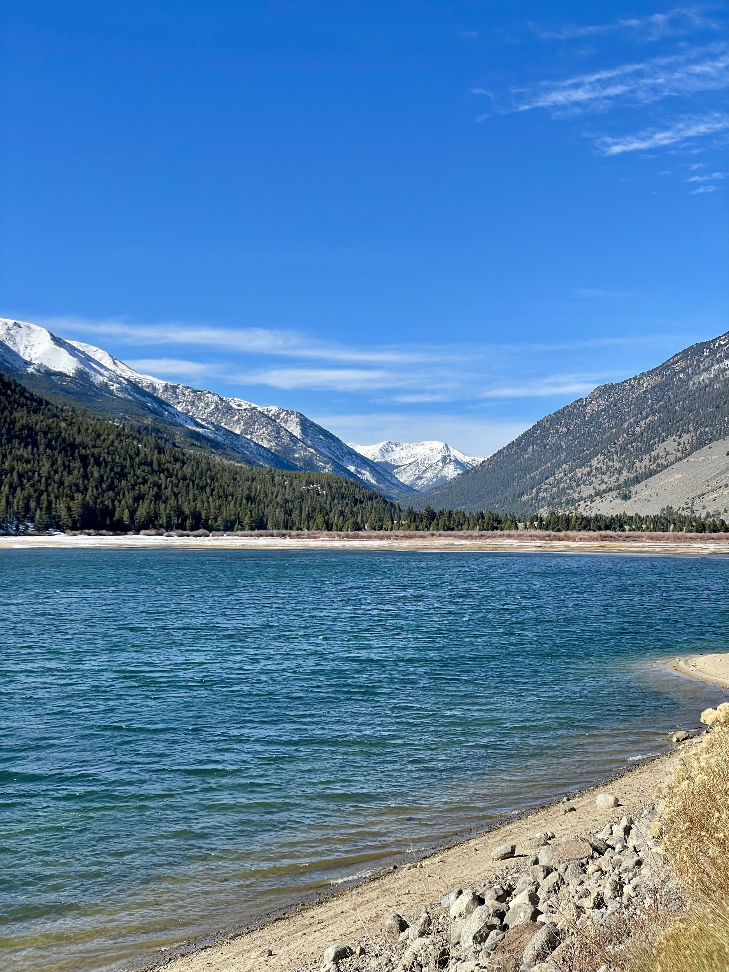 A scenic view of a lake surrounded by snow-capped mountains and dense green forests under a clear blue sky.