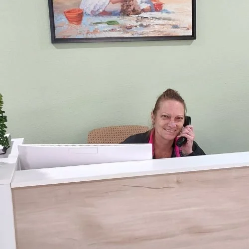 A woman sitting at a reception desk speaking on a phone, with a computer and a small plant nearby, and a landscape painting on the wall behind her.