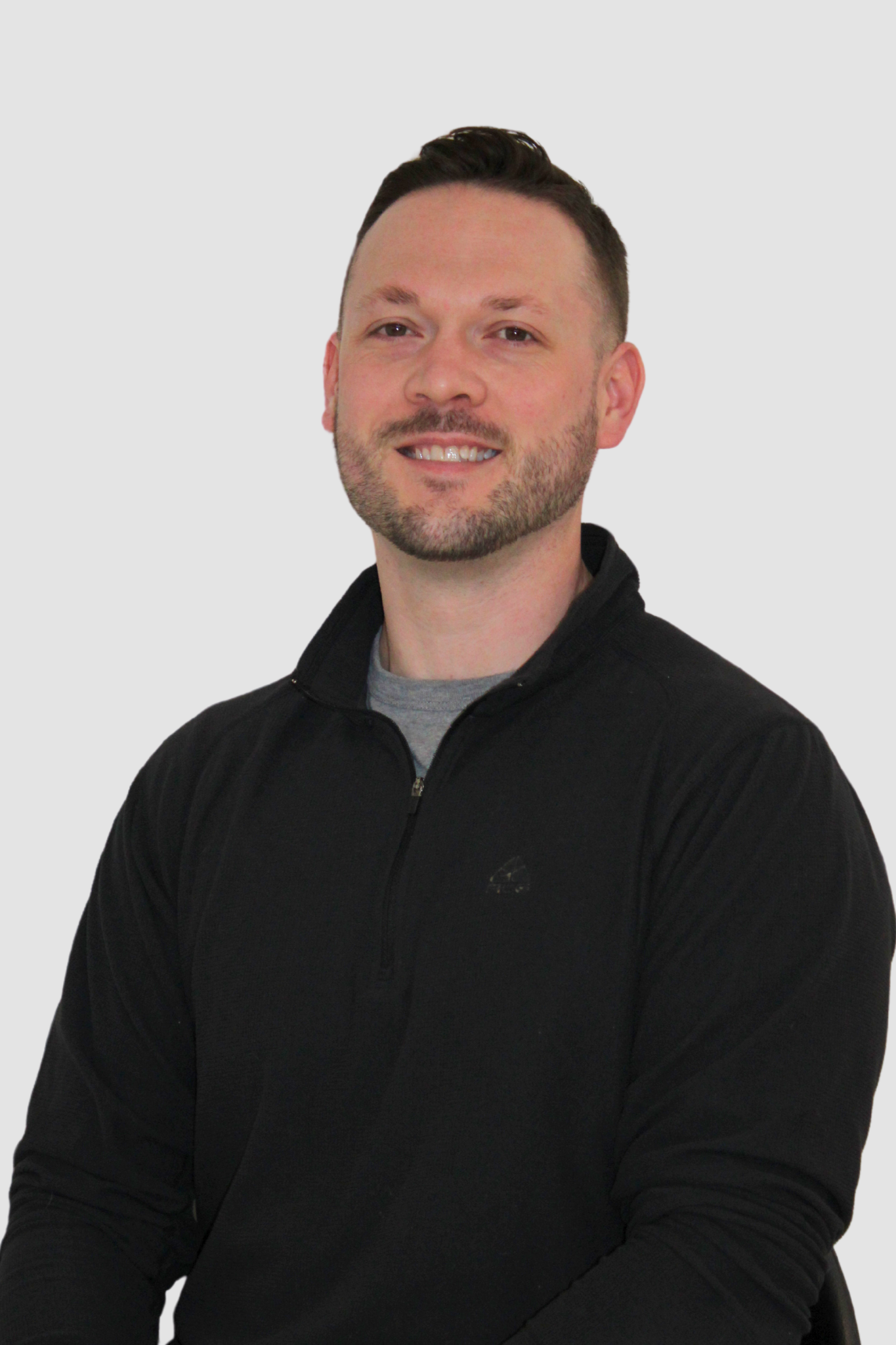 A young man with short dark hair, a beard, and a smile, wearing a black zip-up jacket and a gray T-shirt, sitting against a plain light gray background.
