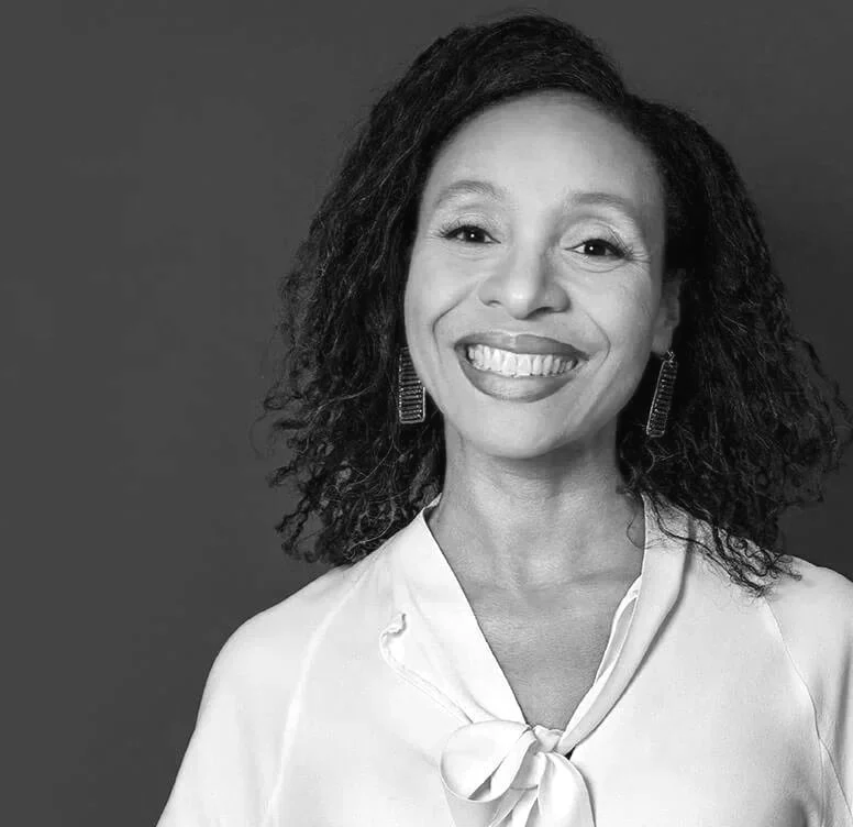 A smiling woman with curly hair and earrings, wearing a blouse with a bow tie in front, in black and white.